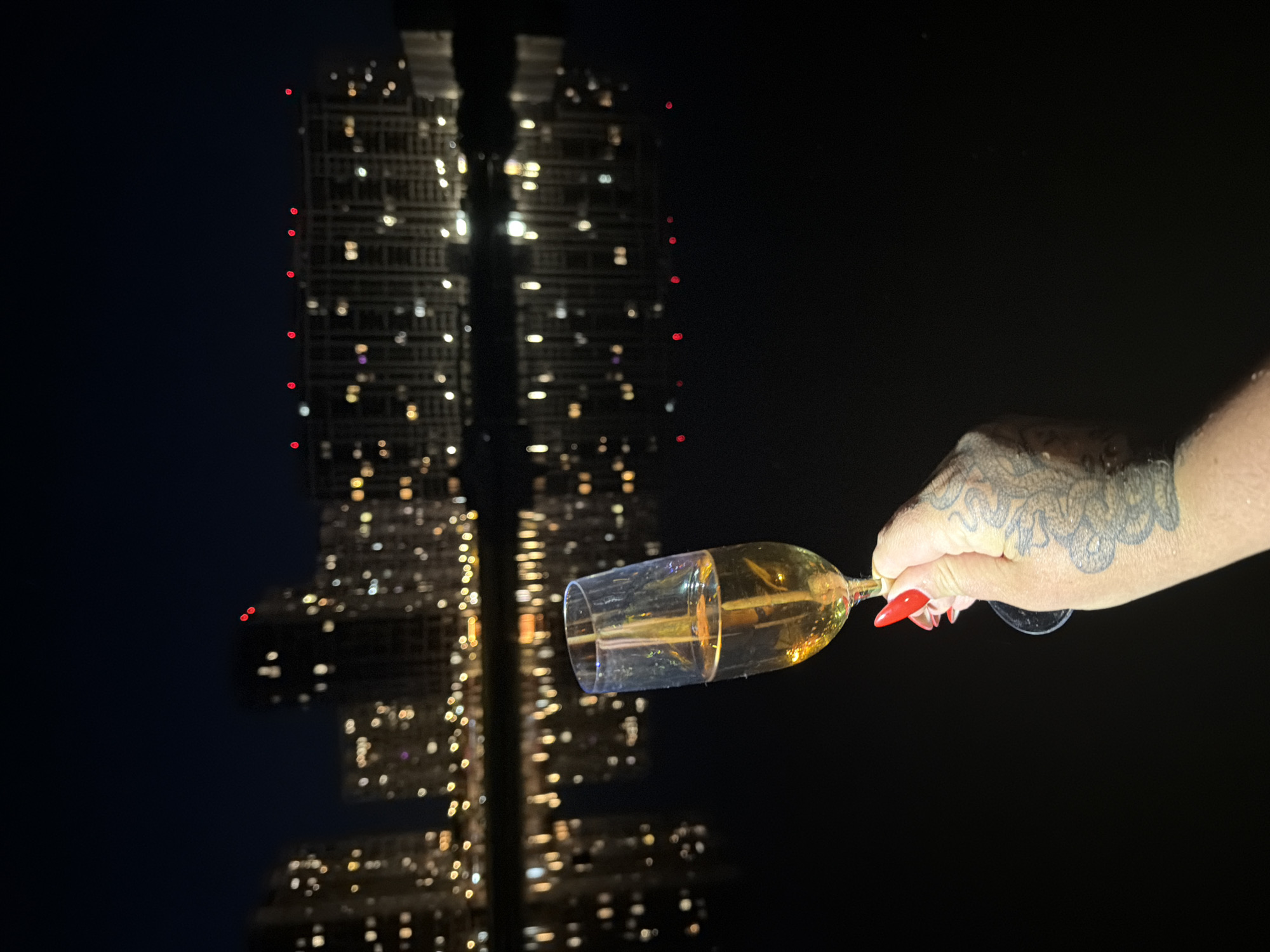 A hand with bright red nail polish and a tattoo on the wrist holds a champagne flute filled with golden liquid. The background is a dark night sky over a body of water reflecting the lights of high-rise buildings. The city lights are blurred, suggesting a long exposure or out-of-focus effect. The scene appears to be set in Kyiv, Ukraine, likely on the Dnipro River embankment, given the characteristic skyline of residential buildings common in the Obolon district. The overall mood is one of celebration or relaxation.