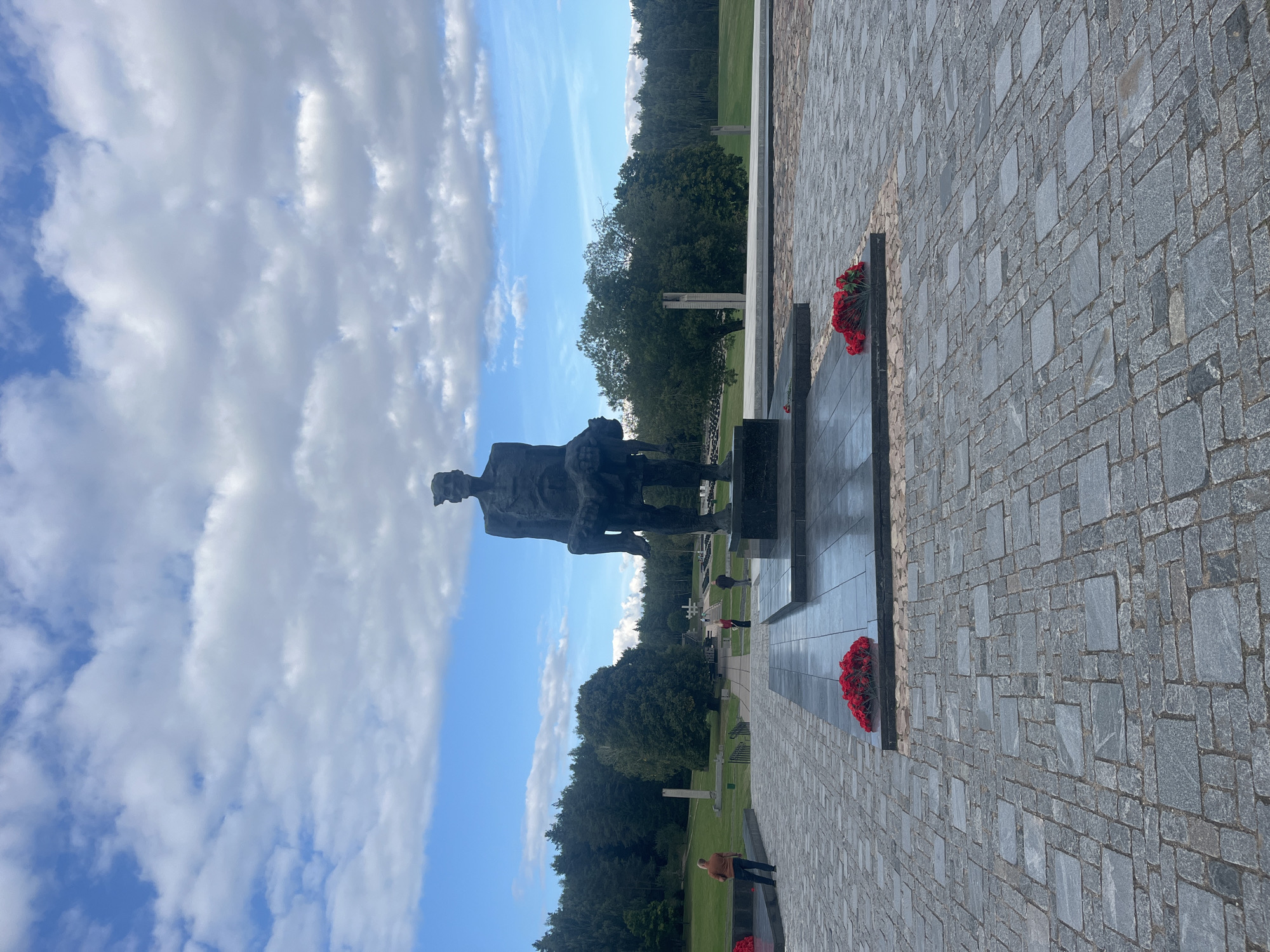 This is a full-shot, eye-level, outdoor photograph of the Khatyn Memorial Complex, located in the village of Каменскі сельскі Савет, Belarus. The main subject is a large, dark bronze statue of a man carrying a child. The statue stands on a raised, black granite platform which is in turn situated on a paved area of grey stone blocks. Red flower wreaths are placed at the base of the statue. In the background, there are several other monuments and structures, including tall, slender pillars and crosses, also surrounded by green grass and trees. A few people are visible in the distance, walking around the complex. The sky is blue with white, fluffy clouds. The overall atmosphere is solemn and memorial-like.