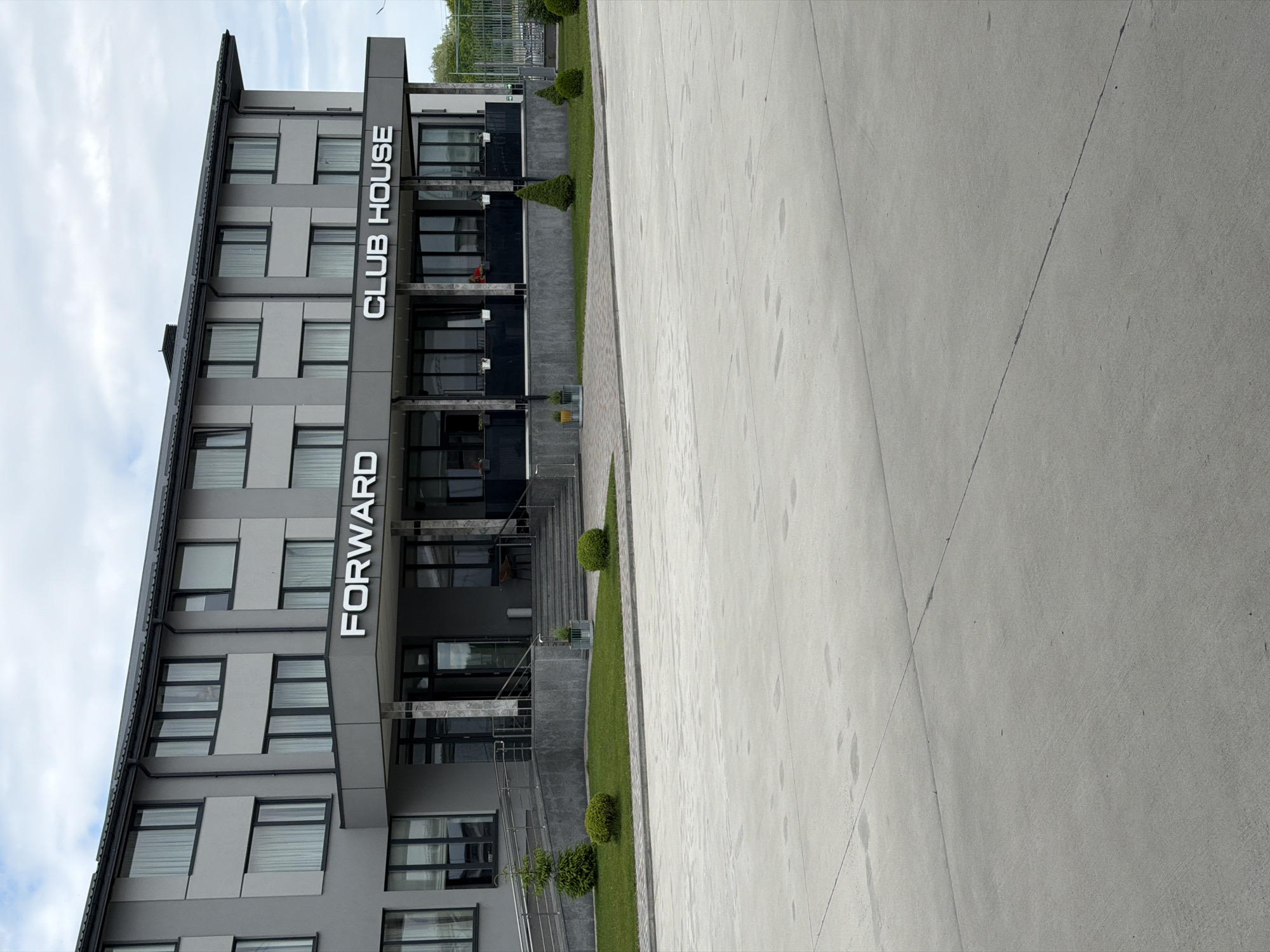 A full, slightly low-angle, eye-level shot captures the exterior of a modern, two-story building in Hnidyn, Ukraine, on a cloudy day. The building has a gray facade with darker gray accents around the windows and the entrance. Large windows with white curtains are visible on both floors. Above the entrance, which features a covered walkway with pillars and glass doors, are the words "FORWARD" and "CLUB HOUSE" in large, white, three-dimensional letters. The entrance is flanked by a gently sloping ramp on the left and stairs on the right, with metal railings. In front of the building, a neatly manicured lawn is dotted with small, conical evergreen trees and spherical shrubs. A paved area, likely a parking lot or plaza, occupies the foreground, with visible concrete seams. The background shows a chain-link fence and some distant trees. There are no people clearly visible in the image, though glimpses of potential figures can be inferred through the glass doors.