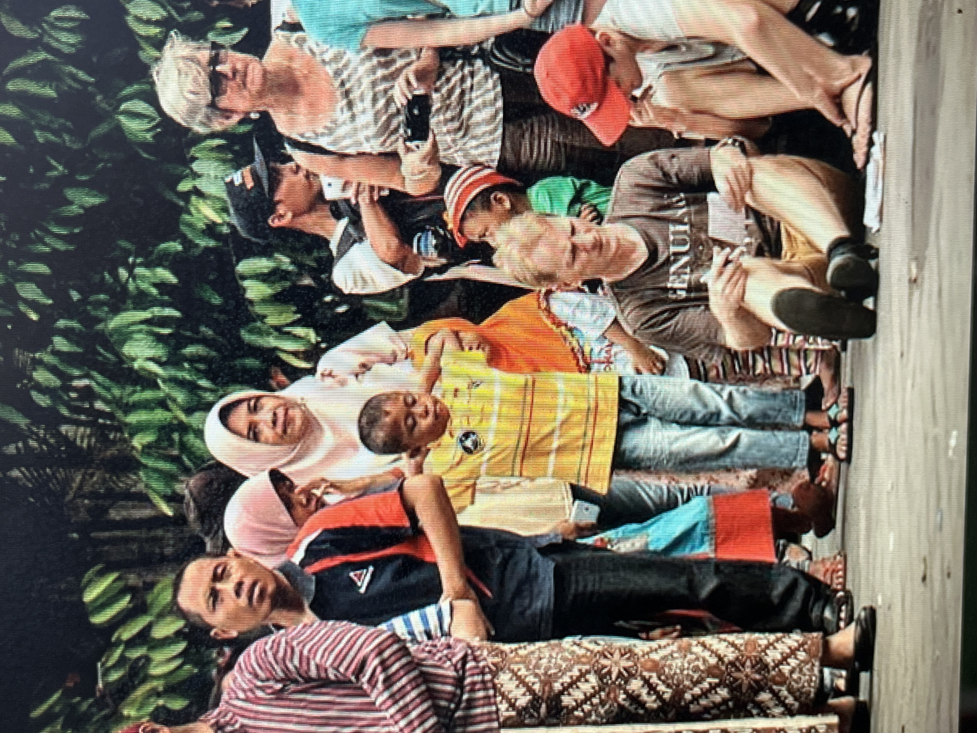 A group of people are standing and sitting on a paved surface, with a backdrop of lush green foliage. The individuals are of diverse ages and ethnicities. One person, wearing a red and white striped shirt and patterned pants, is standing on the left. To their right, a man in a navy and red shirt with a white collar is visible. A young child in a yellow and orange striped shirt is standing in the center, looking down. A woman in a light-colored headscarf is to the left of the child. On the right, a woman with short, gray hair and sunglasses is wearing a patterned top. Beside her, a man in a black baseball cap is holding a smartphone. Two men are seated in the foreground; one in a brown t-shirt is holding a piece of paper, and another to his right is wearing a red baseball cap. The ground appears to be concrete. The overall scene suggests a public gathering or event.