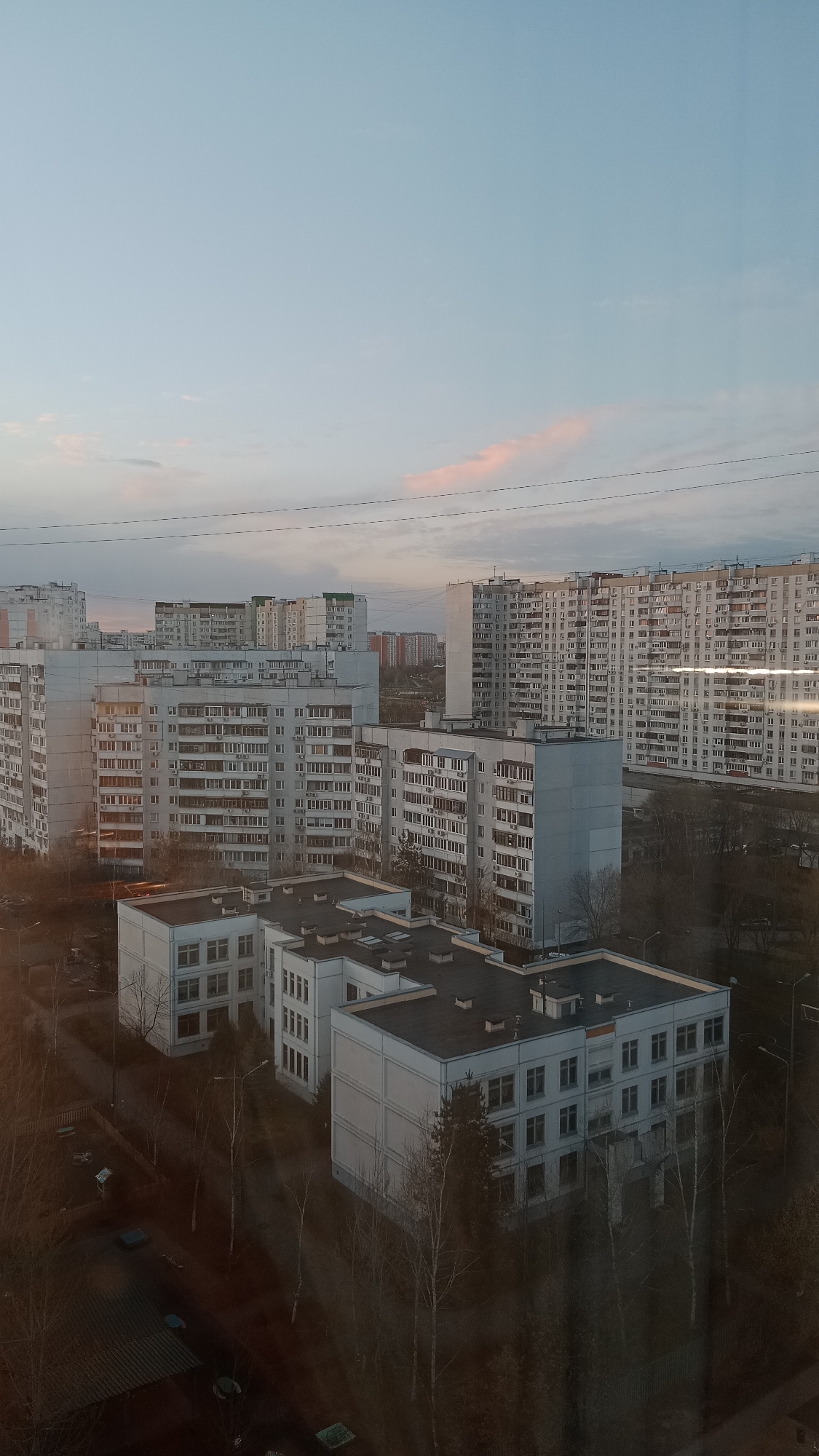 The image shows a view from a high vantage point, likely a window, overlooking a densely populated urban area in Moscow, Russia. The sky is a pale blue with hints of pink, indicating sunrise or sunset. A series of high-rise apartment buildings dominate the horizon, constructed in a utilitarian Soviet-era style, characterized by repetitive rows of windows and prefabricated concrete panels. Closer to the viewer, there are several lower buildings, possibly schools or administrative offices, with flat roofs and similar architectural features. Trees surround these lower buildings, most of which are bare, suggesting late autumn or winter. Some cars are visible in parking areas. Electrical wires stretch across the sky. The overall impression is of a cold, urban landscape. The image has a slightly blurred quality, possibly due to reflections on the window or atmospheric haze. There are no discernible people in the image.