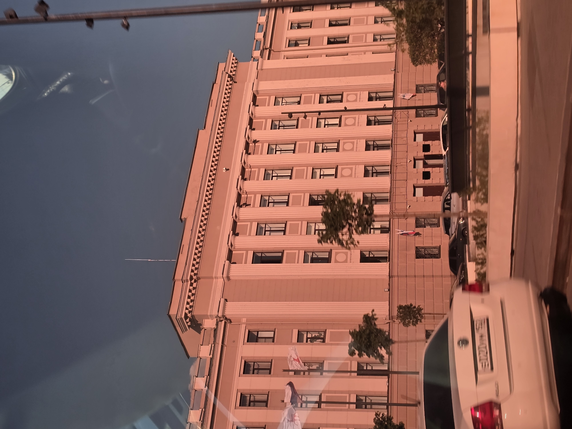A long, pink building with a classical facade, featuring columns and multiple windows, is illuminated by the warm light of a setting sun. Two flags, one red and white and another with a white background and red symbols, are visible on flagpoles in front of the building. Several trees are planted along the facade. In the foreground, a white car is partially visible, with its license plate reading "E202MM 152". Other cars and a dark road are also present. The sky is a deep blue, with a hint of clouds. The scene appears to be in Nizhny Novgorod, Russia, possibly near the Kremlin.
