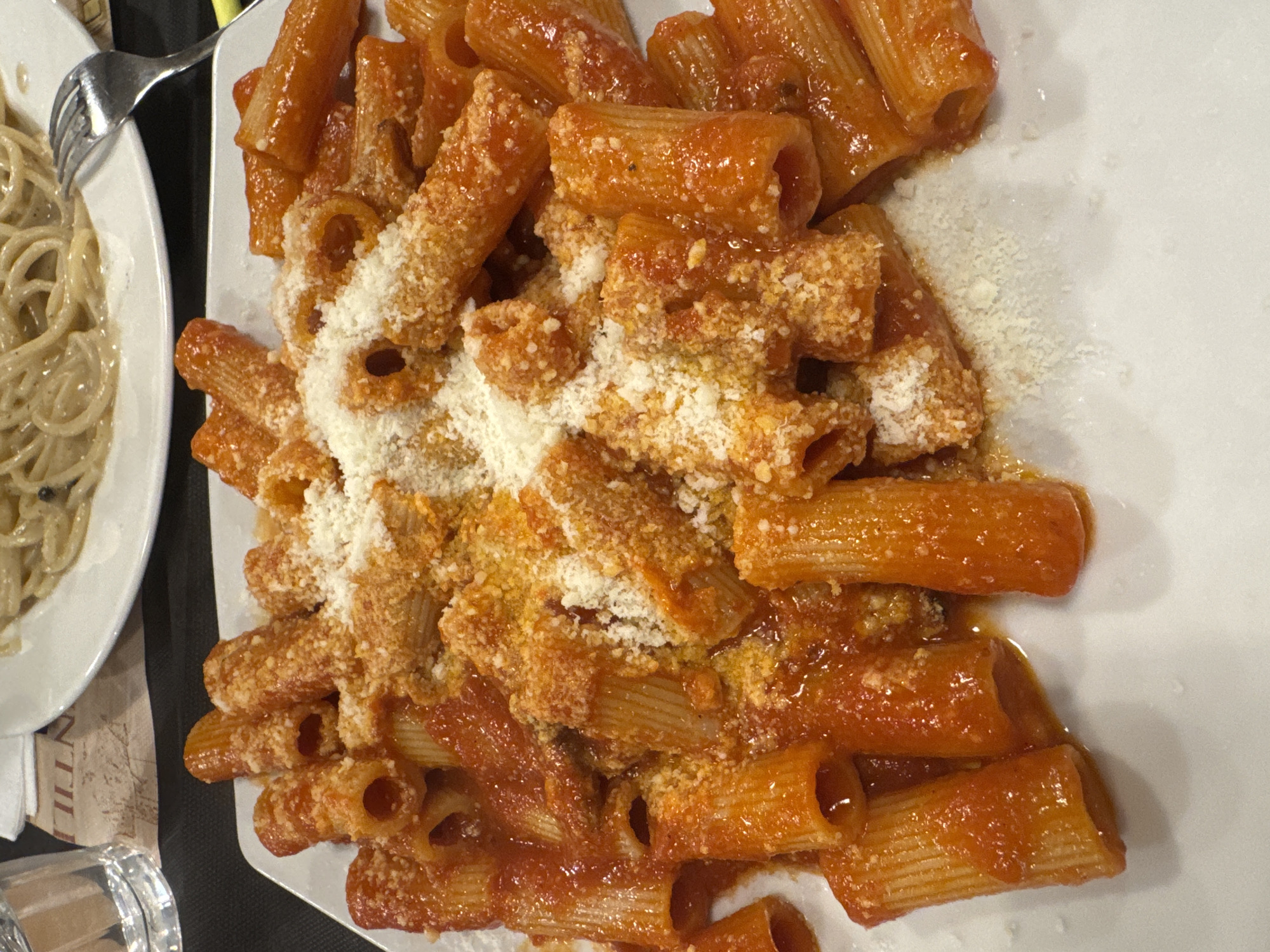 The image displays a dining scene within an Italian restaurant in Rome, Italy. In the foreground, a white rectangular plate is centrally positioned, laden with rigatoni pasta. The pasta is coated in a red tomato-based sauce and generously topped with finely grated white cheese. In the blurred background, a partial view of a round white plate reveals spaghetti covered in a light-colored creamy sauce, with a silver metal fork resting beside it. A clear glass filled with water is visible on the far left. The table surface is dark, and a patterned paper placemat or menu is visible under the background plate, displaying the partial uppercase text "NTILLI". No individuals are present; the scene focuses exclusively on the prepared food.