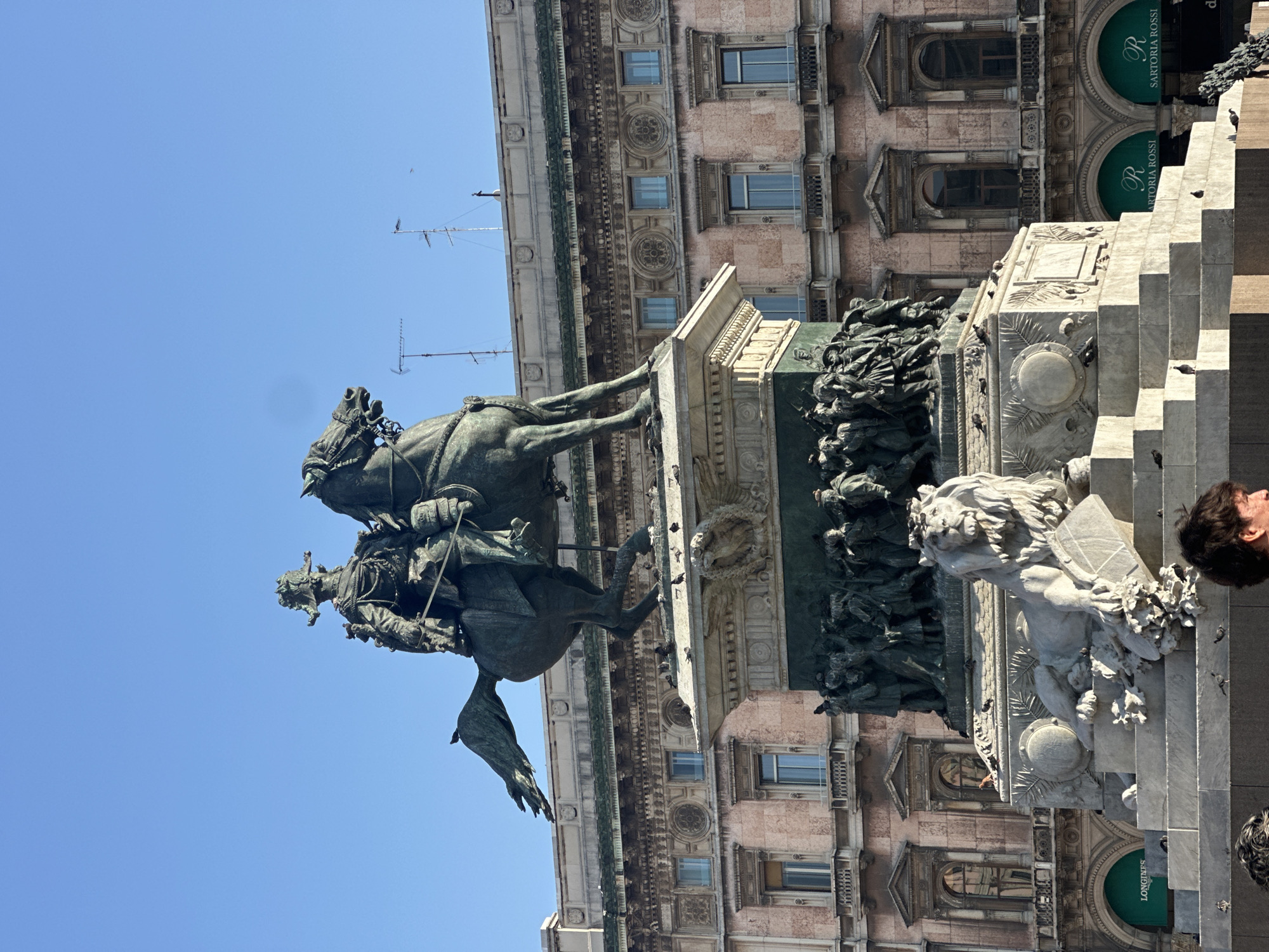 This is a medium shot of the monument to Vittorio Emanuele II in Piazza Duomo, Milan, Italy. The monument, a large bronze equestrian statue atop a stone pedestal, is centrally framed against a clear blue sky. The statue depicts a man in military attire on horseback, with a group of figures at the base. In front of the pedestal, a stone lion reclines on a stepped platform. The background features a historic building with arched windows and decorative elements. Several pigeons are visible on the monument and the surrounding platform. The lower portion of the image shows the heads of two individuals, partially obscuring the monument's base. Green awnings with the logo "SARTORIA ROSSI" are visible on the ground level of the building in the background.