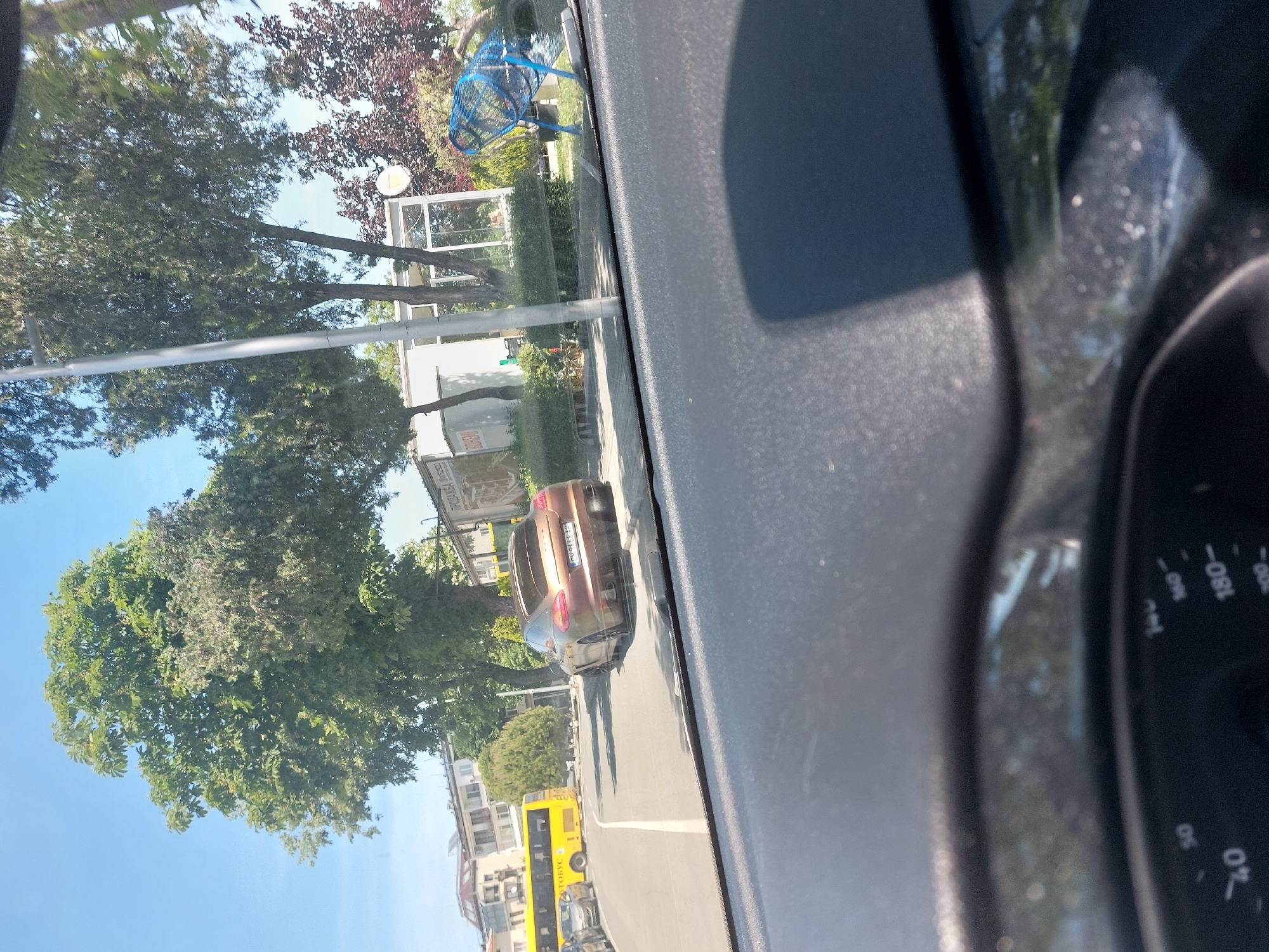 The image shows a street scene from inside a car, likely in Tsarevo, Bulgaria. The camera is looking out the windshield, capturing a view of the road ahead, a parked car, and trees lining the street. A yellow bus is visible in the distance. A park with a blue play structure is visible in the background. The dashboard of the car is visible in the foreground, including the speedometer.