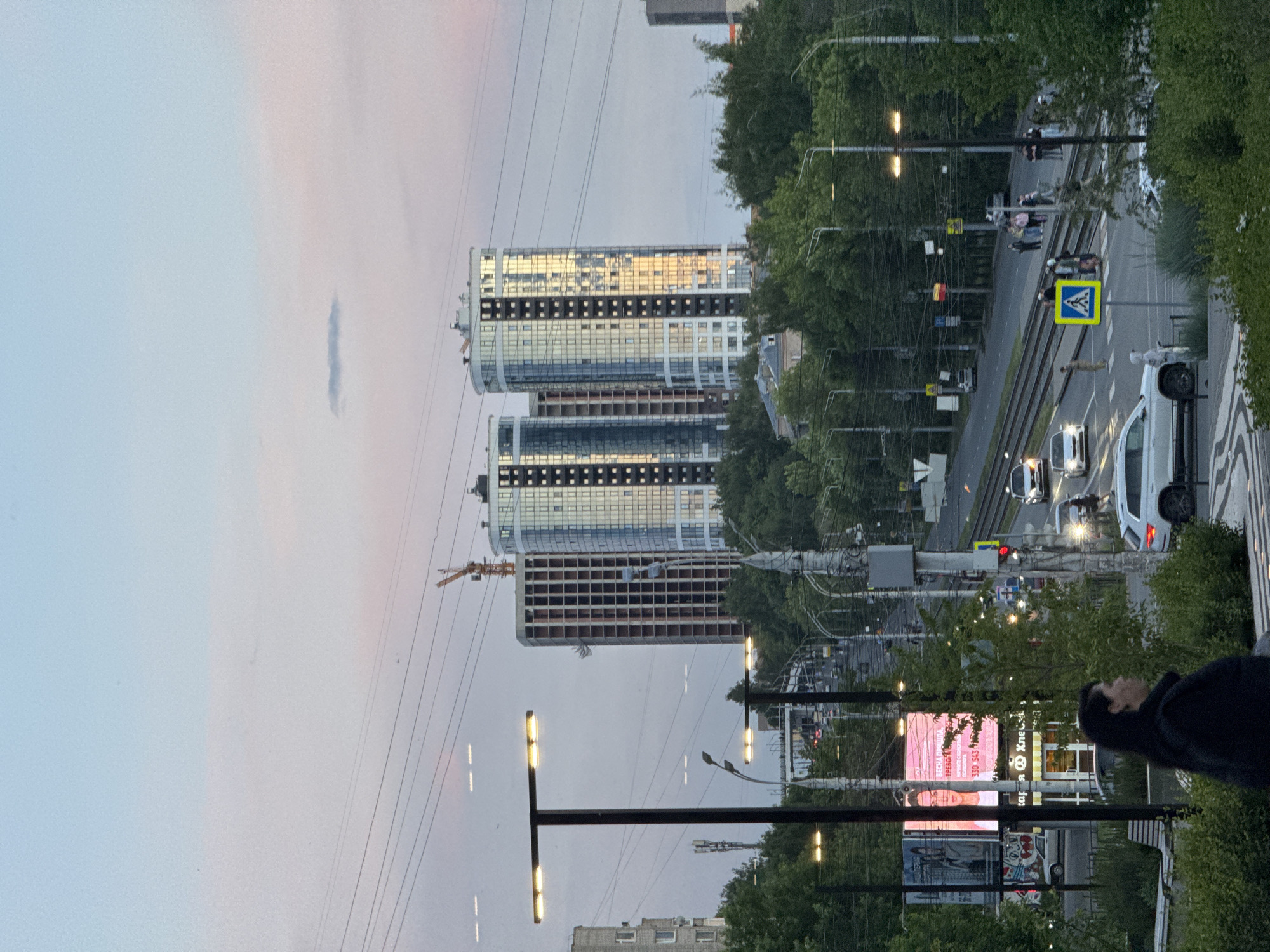 The image depicts an urban street scene in Izhevsk, Russia, captured during twilight, likely early evening. The sky, a gradient from light blue to a soft pink, suggests either sunrise or sunset. A thin, elongated cloud floats in the sky.
In the background, two modern high-rise buildings stand prominently. The taller building features extensive glass facades reflecting the ambient light. The shorter building appears to be under construction, with exposed frameworks on its lower floors, and a construction crane is visible to its left.
The mid-ground is dominated by a multi-lane road with tram tracks running through its center. Several vehicles, including a white van or SUV in the foreground, are present on the road. A clear blue pedestrian crossing sign marks a crosswalk where a few pedestrians are visible. Trees line the street, providing greenery. Numerous streetlights are active, casting a warm glow, and power or tram lines crisscross the upper part of the frame.
In the right foreground, a person with dark hair, wearing dark clothing, is partially visible, partially obscured by a streetlight pole. Further down the street, various commercial signs are present. A large billboard prominently displays Cyrillic text, which translates to "Spring demands" ("ВЕСНА ТРЕБУЕТ") and "Demand yours" ("ТРЕБУЙ СВОЁ"), along with phone numbers "330 545" and "56-02-03". Another partial sign reads "Хлеб" (Bread), likely indicating a bakery or shop. The scene portrays a typical busy city environment during the transition from day to night.