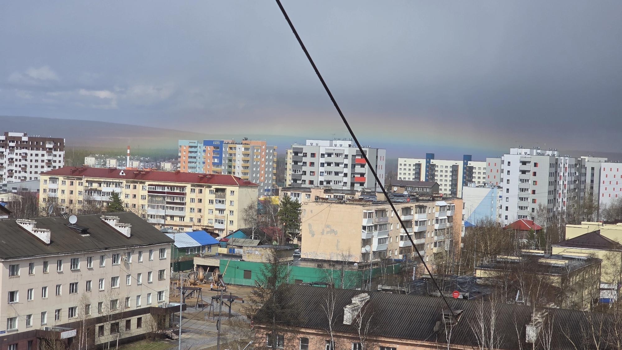 This image presents a cityscape in Lensk, Russia, observed from an elevated vantage point. Predominantly, the frame is filled with multi-story residential buildings, exhibiting a variety of architectural styles and paint schemes ranging from beige to blue, orange, gray, and white. Some buildings have red roofs. The buildings are situated among trees.
In the backdrop, a faint rainbow arcs across the skyline under a cloudy sky. A black power line cuts diagonally across the upper portion of the frame. At the forefront, building rooftops dominate the immediate foreground. Some trees have lost their leaves, indicating a time of year like autumn or early spring. No people are immediately visible in the frame. The overall impression is of an urban scene in a northern environment.