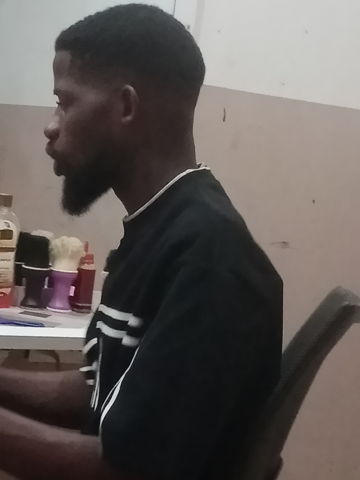 The image is an indoor, medium shot of a Black male sitting in a chair, viewed from the right side. He has dark skin, short black hair, and a beard. He is wearing a black t-shirt with white stripes on the sleeves. A white-lined shelf is in the foreground, holding several items including a bottle with a yellow cap, a purple cup with a white object inside, and a red bottle. The background is a plain, light-colored wall with some subtle texture. The lighting appears to be artificial. The scene is likely in a personal grooming space or barber shop. The image location is stated to be Jalingo, Nigeria.