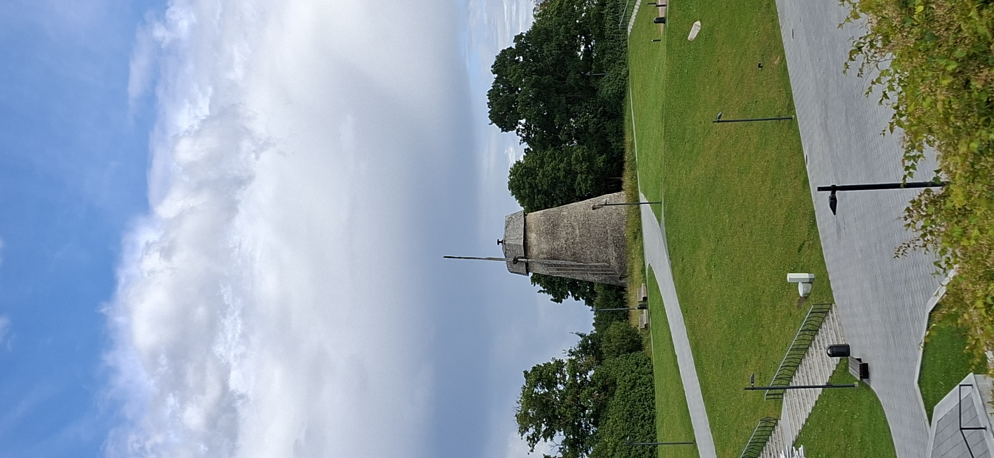 The image depicts a scenic view of a traditional windmill standing in a grassy park area. The windmill is the central focus, constructed of stone with a conical shape and a visible cap. It is surrounded by lush green trees and well-maintained pathways. The sky above is a mix of blue and dramatic grey clouds, suggesting dynamic weather conditions.
The scene is identifiable as being located in the city of Rakvere, Estonia. Specifically, this is the area around the Rakvere Castle, where this preserved windmill is a notable landmark. The pathways, benches, and lampposts indicate a public park designed for visitors. There are no people visible in the image. The overall impression is one of historical preservation within a natural, landscaped setting.
