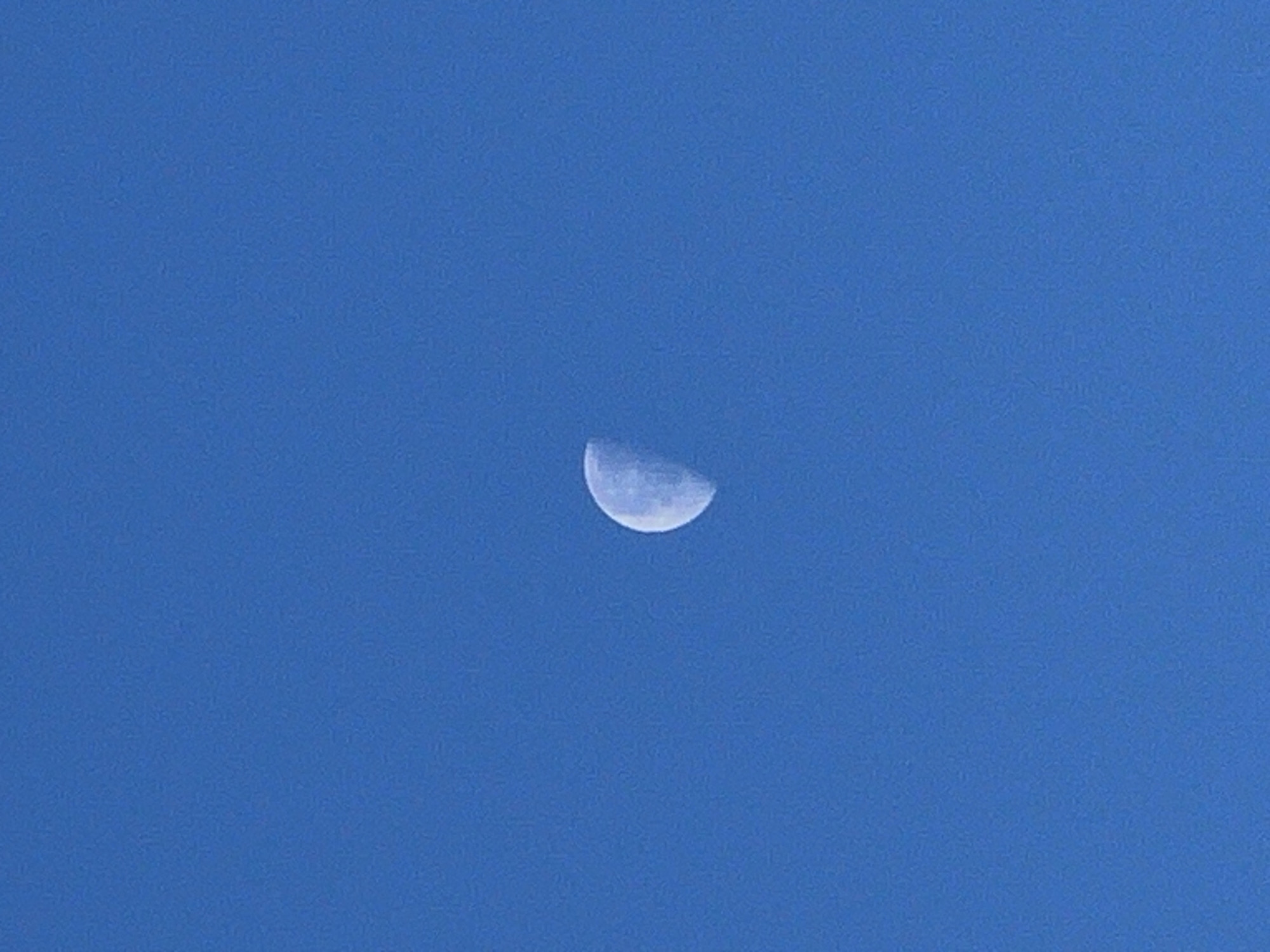 A crescent moon is visible in a clear, bright blue sky. The moon is illuminated on its left side, with the right side appearing dark. The image is framed to focus on the moon against the expanse of the sky.