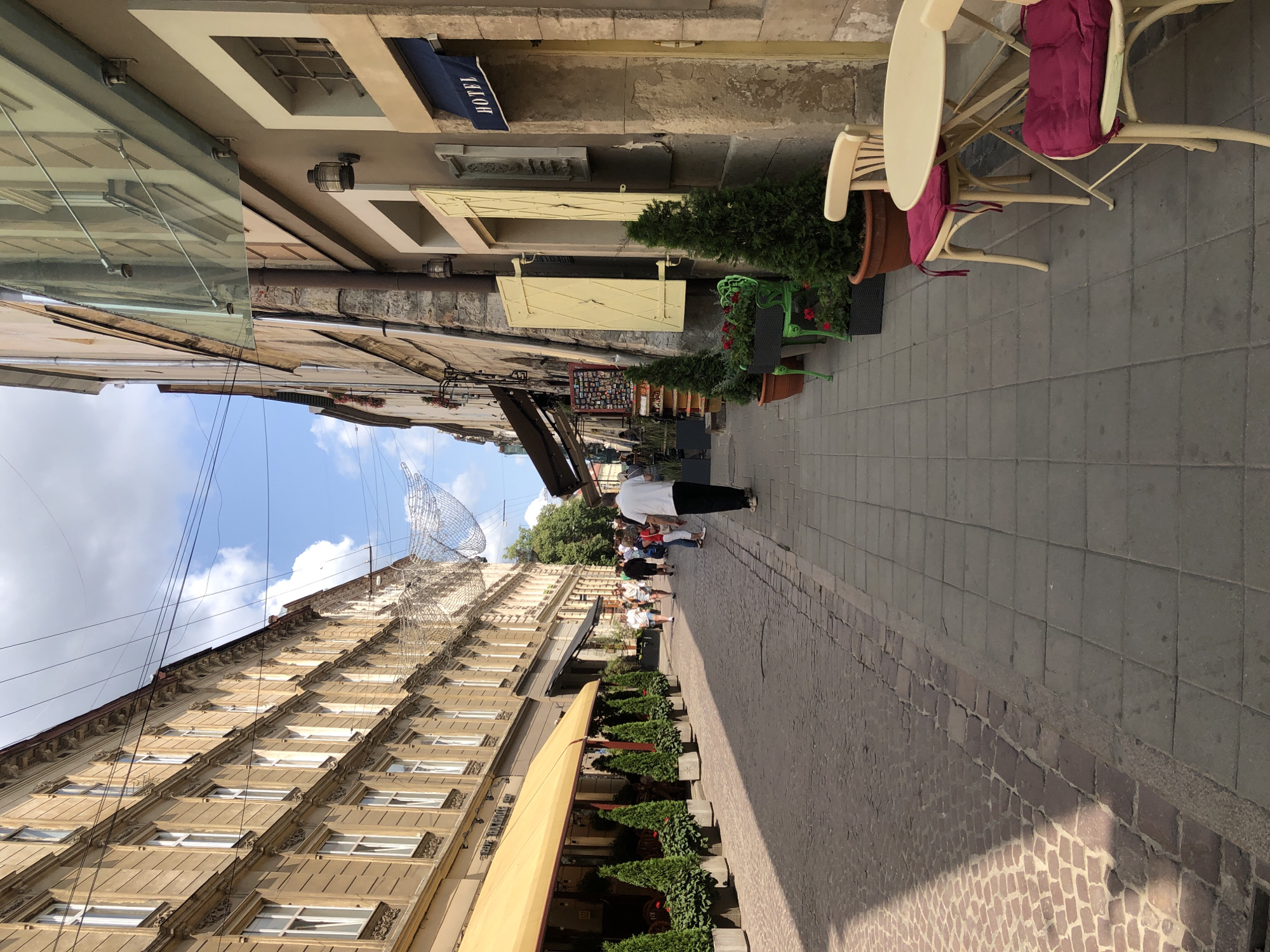This is a ground-level, eye-level, long shot of a European city street in Lviv, Ukraine. The street is paved with cobblestones and has sidewalks on either side. The buildings lining the street are old and ornate, with multiple stories and large windows. Several cafes and shops are visible along the street. There are a few people walking on the street, but it is not crowded. The sky is blue with some clouds. The image was taken on a sunny day.