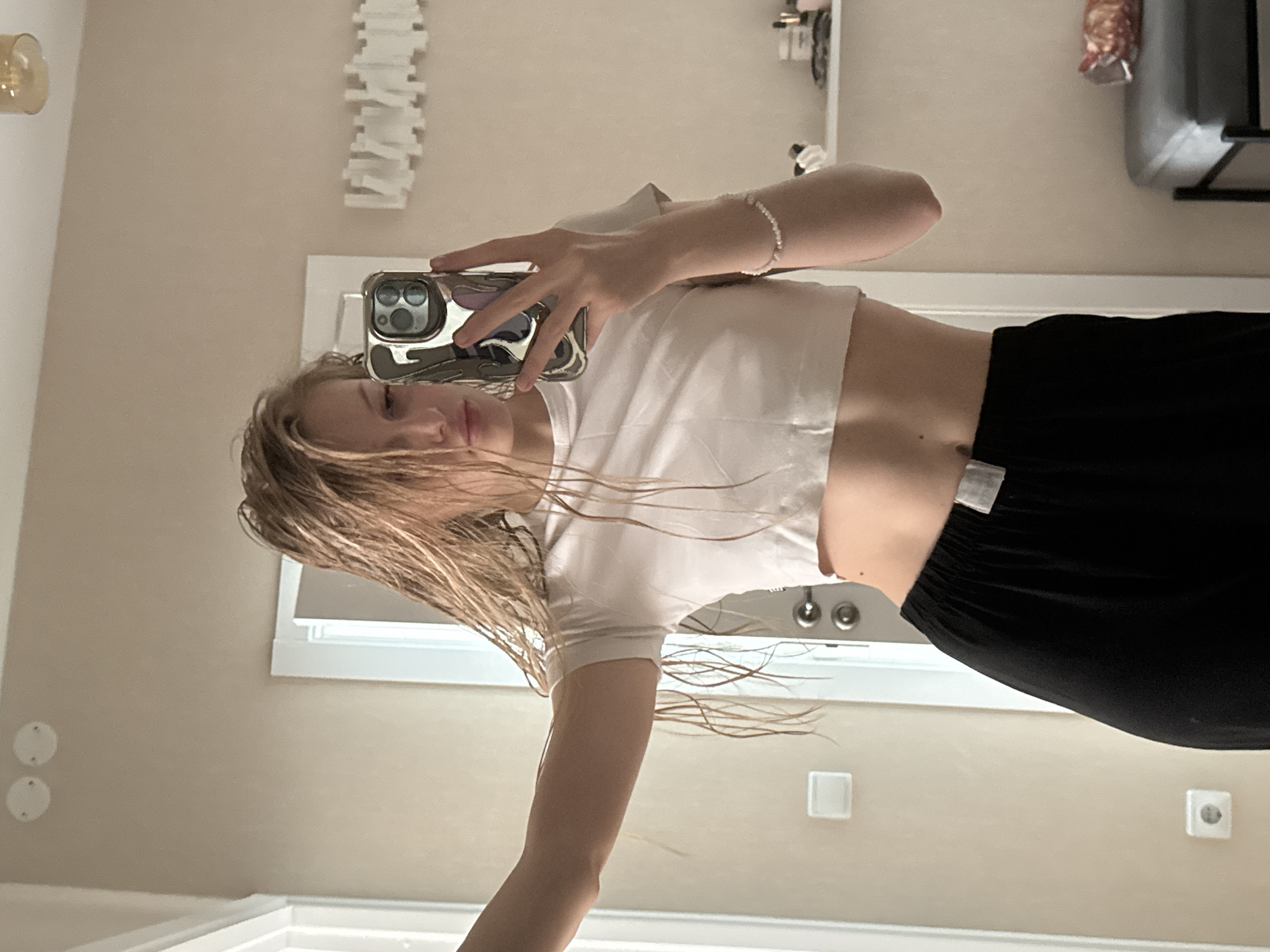 A young person with wet blonde hair is taking a selfie in a bathroom mirror. The individual is wearing a white cropped t-shirt and black pants. The bathroom has a light beige wall, a white window frame, a modern white light fixture, and electrical outlets. The city of Perm, Russia, is not discernible from the provided image.