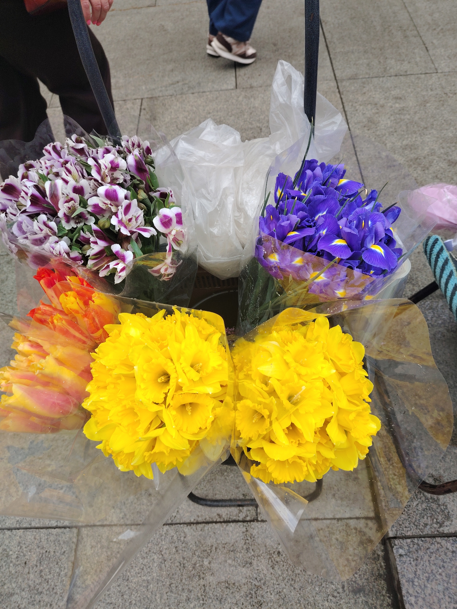 The image shows a collection of four flower bouquets in individual plastic wraps arranged in a container, likely for sale. The bouquets consist of: purple and white alstroemeria, blue irises, orange tulips, and yellow daffodils. The background shows a paved area suggesting a street. Portions of two people's legs and shoes are visible, indicating the presence of individuals nearby. The container holding the flowers is partially obscured, but appears to be part of a larger structure, possibly a bicycle or cart. There is no information to indicate the location is in Moscow, Russia.
