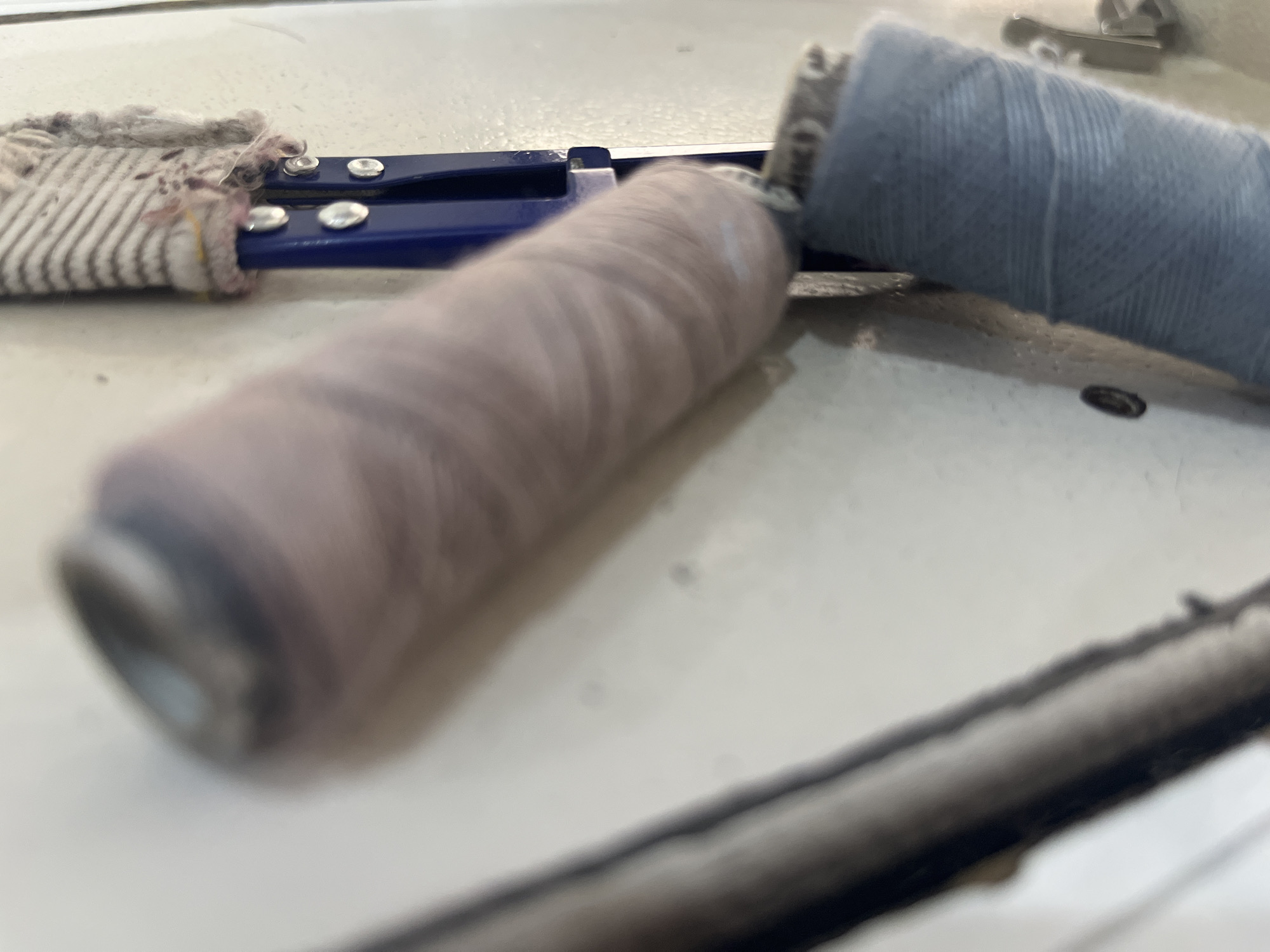 The image shows two spools of thread, one pale pink and one light blue, resting on a white surface, possibly a table or countertop. A blue clamp-like object is visible under the spools. A piece of striped fabric, possibly a cloth or rag, is attached to the clamp. The scene appears to be an indoor setting, with no specific landmarks to suggest a city location. The lighting is bright and diffused, casting a slight shadow.