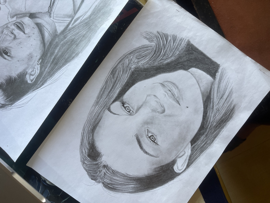 Two grayscale pencil portrait drawings are displayed on white paper in an unspecified indoor setting in Jos, Nigeria. The foreground drawing, angled towards the bottom left, is a head-and-shoulders portrait of a woman with long, dark, wavy hair, parted on her left side, and a mole below the right corner of her mouth. Her face is turned slightly to her right, with her gaze directed towards the viewer. Behind and to the right, partially obscured by a dark rectangular object, is a second pencil portrait depicting the head and upper chest of another individual, possibly a woman, with long hair pulled back, an ear visible with a 'paperclip'-shaped earring, and facial features suggesting freckles or skin texture. Both drawings exhibit detailed shading and appear to be realistic representations. The papers show minor creases, and they rest on a dark, reflective surface, suggesting a table or desk.