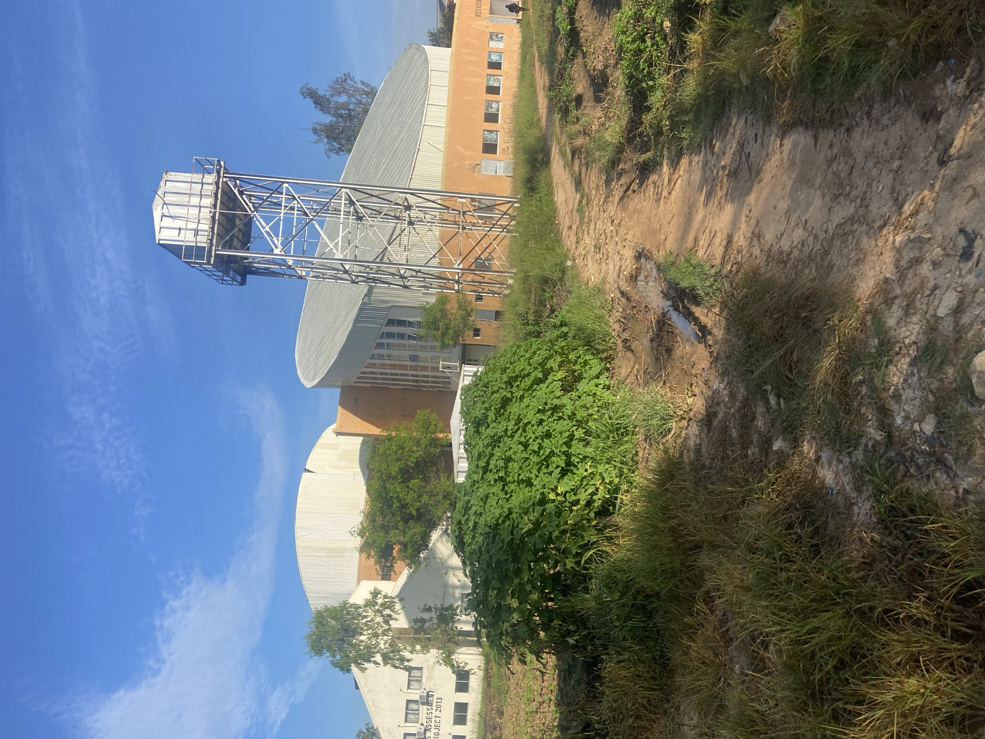 The image depicts a large, modern building with a distinctive curved roof structure, likely a community hall or sports facility. The building's facade is a mix of light-colored panels and orange-colored sections. To the right of the main structure stands a tall, metallic water tower. In the foreground, a dirt path winds through overgrown grass and vegetation, with some evidence of puddles suggesting recent rain. The sky is bright blue with scattered white clouds. The scene appears to be located in a semi-rural or developing area, with the building representing a significant infrastructure project in what is identified as the city of Rafin Par, Nigeria. A lone figure is visible walking on the path near the building's entrance.