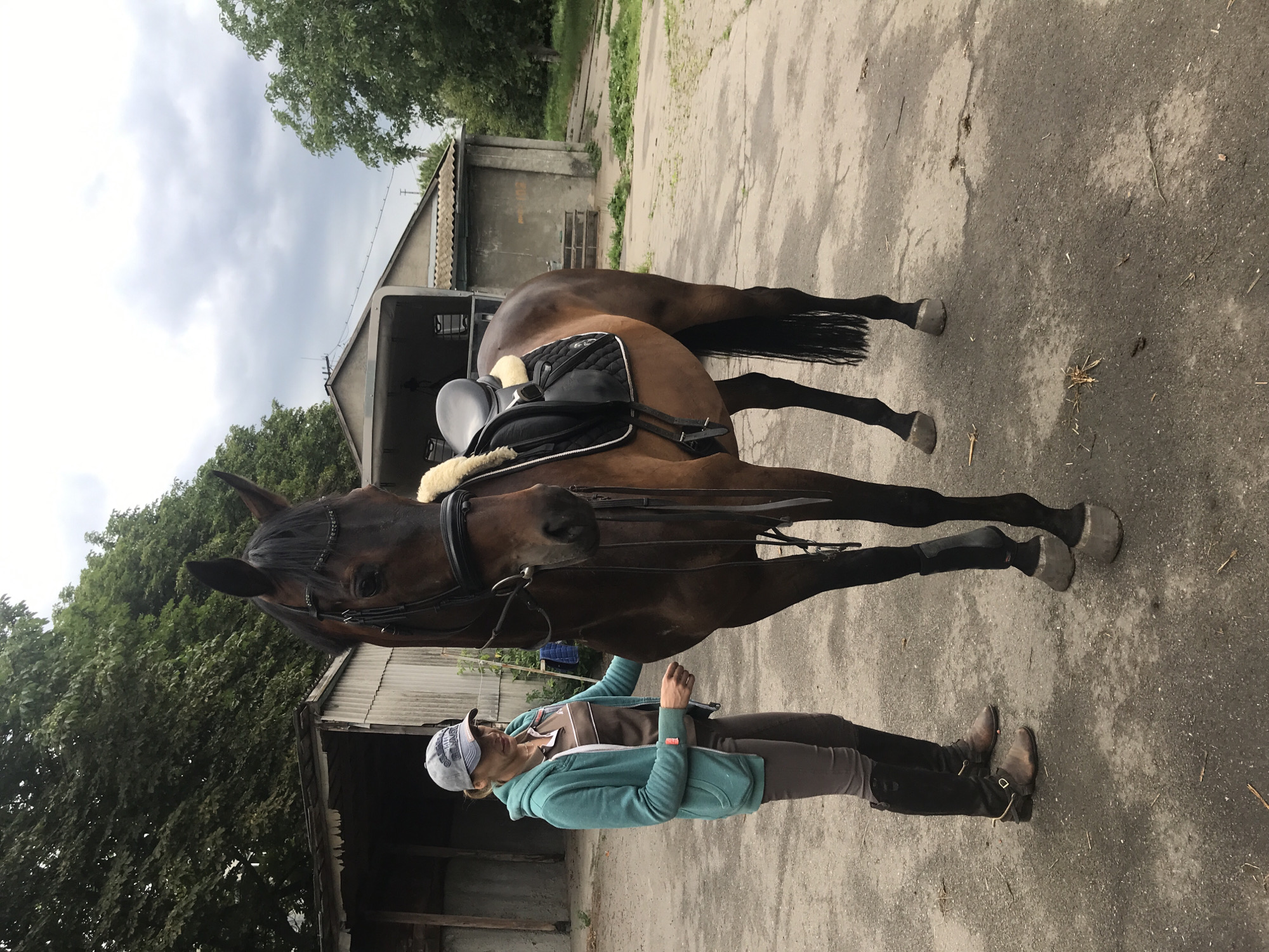 A woman is standing next to a brown horse, which is saddled and bridled. The woman is wearing a teal hooded jacket, brown pants, and tall black boots. She has on a light-colored baseball cap. The horse is a dark brown color, with a black mane and tail, and is wearing a black saddle and bridle. The scene appears to be outdoors, possibly at a stable or riding school, with a building in the background. The ground is paved with concrete. The image is taken from a slightly elevated angle. The lighting suggests it is daytime, with overcast skies.