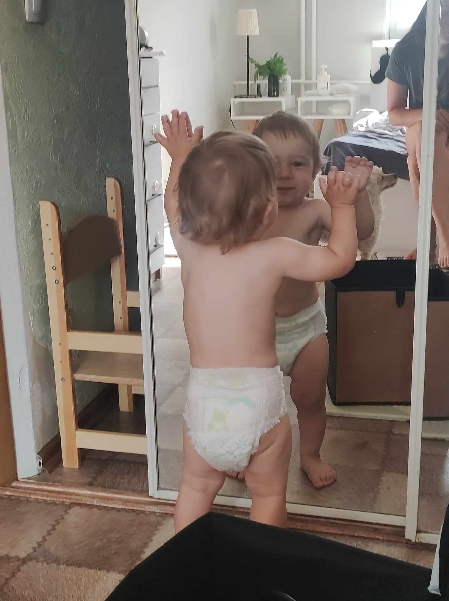 The image depicts a toddler in a diaper standing in front of a full-length mirror. The toddler is facing the mirror with their hands raised as if touching the mirror's surface. The toddler's reflection is visible in the mirror. The background reveals a room setting with furniture, including a child's learning tower and a white cabinet. Reflected in the mirror is an adult sitting on a bed, a dog-like figure, a black trunk, white shelving, a lamp, and plants. The location is an indoor residential setting, presumably in a home. There is no explicit evidence that indicates the image was taken in the city of Chaikovsky, Russia.
