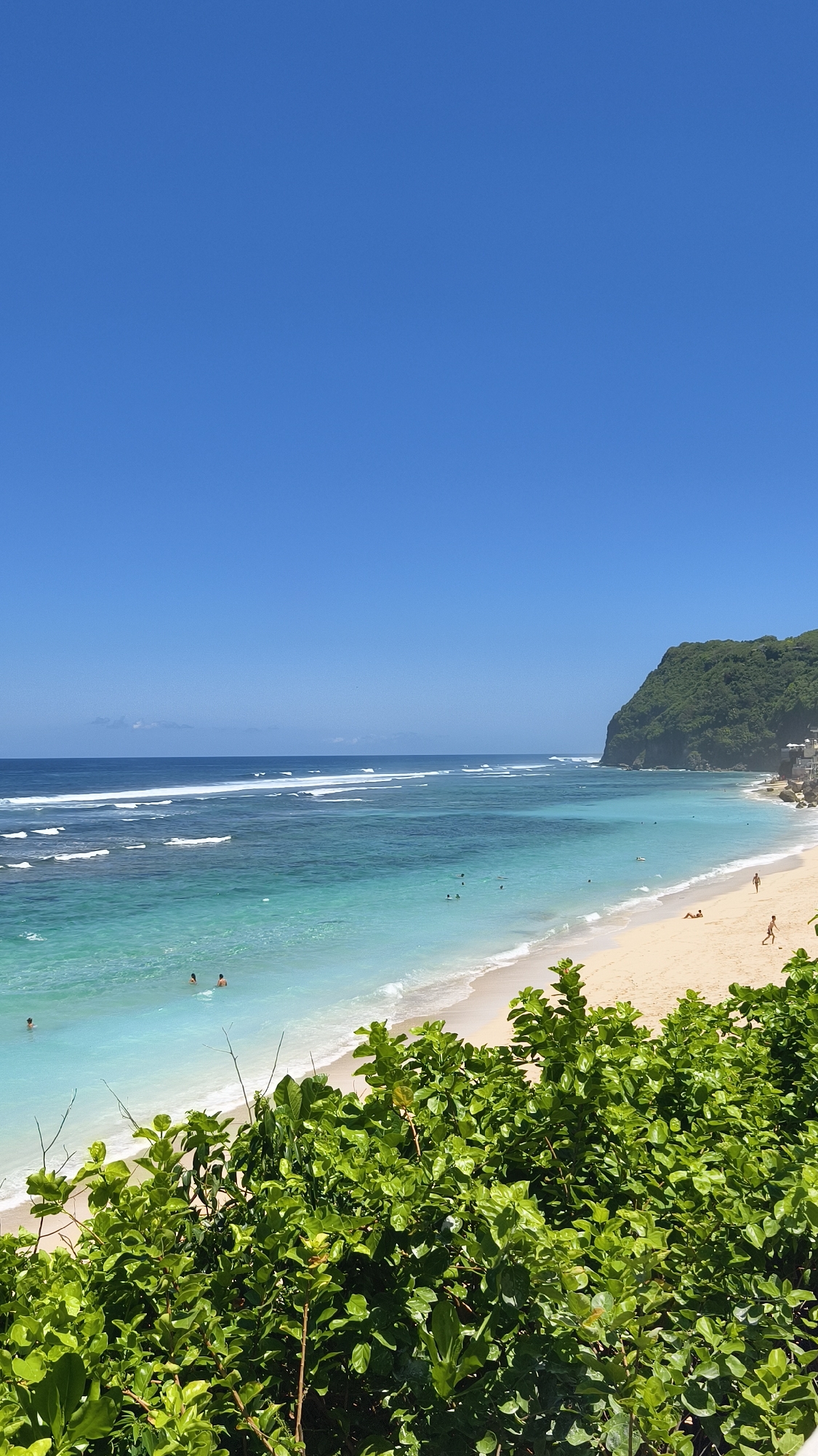 The image captures a coastal scene in Kuta Selatan, Indonesia. The scene includes a sandy beach with several people scattered along the shoreline and in the water. The ocean transitions from a clear turquoise near the shore to deeper blue hues further out, with visible waves. A small, forested hill or headland is visible at the edge of the beach in the distance. The sky is a clear, bright blue. In the foreground, a dense patch of green vegetation obscures the lower portion of the view.