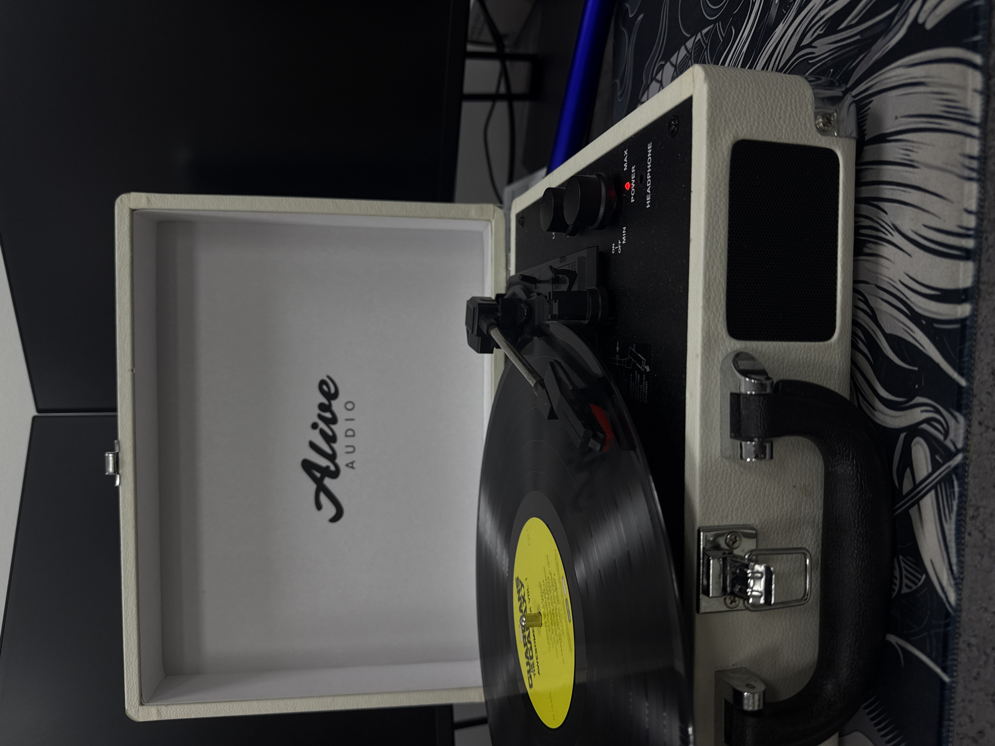 The image shows an open suitcase-style record player, with a vinyl record on the turntable. The record player is white with a black control panel and a black handle on the side. The inside of the lid is also white, with the text "Alive AUDIO" printed in black. The record on the turntable has a yellow label with text. The record player is sitting on a patterned surface with black, white, and grey designs. Part of a black monitor is visible in the background. There is no evidence to suggest the image was taken in Chaikovsky, Russia.