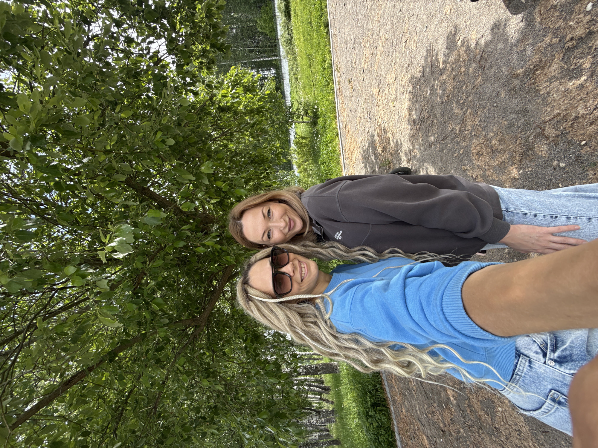 Two women stand on a dirt path in Заволжье, Russia. The woman in the foreground wears a blue sweatshirt, jeans, and sunglasses, with blonde braided hair. The other woman wears a dark gray sweatshirt and jeans. A tree with green leaves occupies the left side of the frame. A grassy area is behind the tree. A dirt path leads into the background.