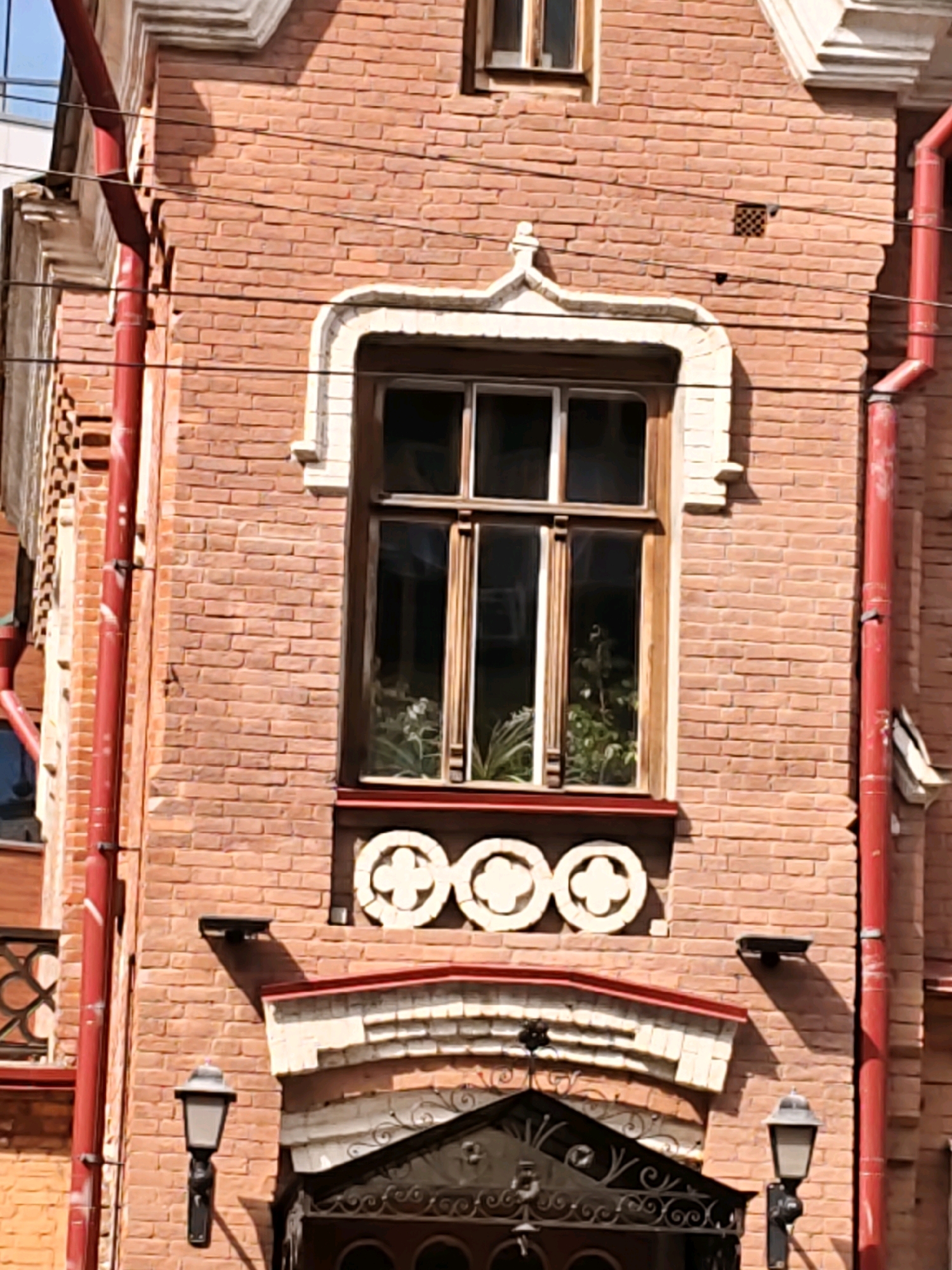 The image shows a close-up view of the facade of a brick building. The building has a red brick exterior with decorative white trim around a large window. The window has wooden frames and is divided into multiple panes. Inside the window, there are plants. Below the window, there are three circular decorative elements with cross-like designs. There are also two outdoor lamps attached to the building's facade. Red downspouts are visible on the right and left sides of the building. The overall style suggests it is likely an older building, possibly of architectural interest.
This building is located at Krasnoarmeyskaya Street, 37, Rostov-on-Don, Russia. This specific building is the former mansion of merchant F.V. Panin.