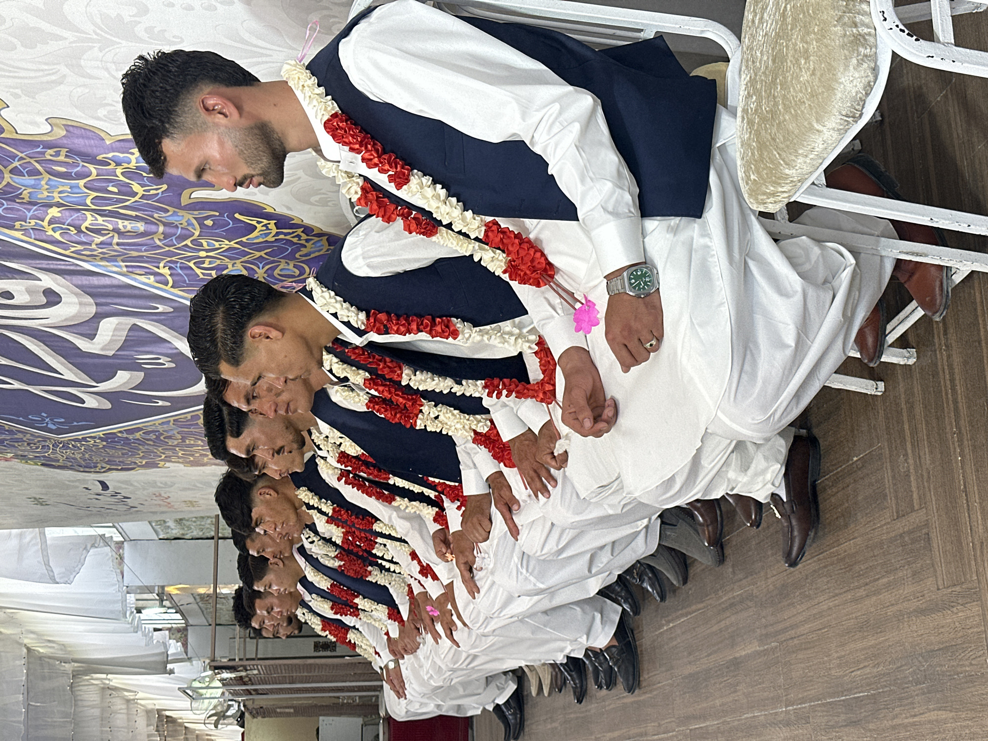 The image shows a row of men seated in chairs, possibly at a formal event. They are all dressed in similar attire consisting of white shirts and pants with dark blue vests, and each man is wearing a garland of red and white flowers around their neck. The men all appear to be looking down or towards the side. The setting appears to be indoors, with a patterned ceiling visible in the background. The presence of similar clothing and garlands suggests a shared role or participation in the same event. The location is Quetta City Tehsil, Pakistan.