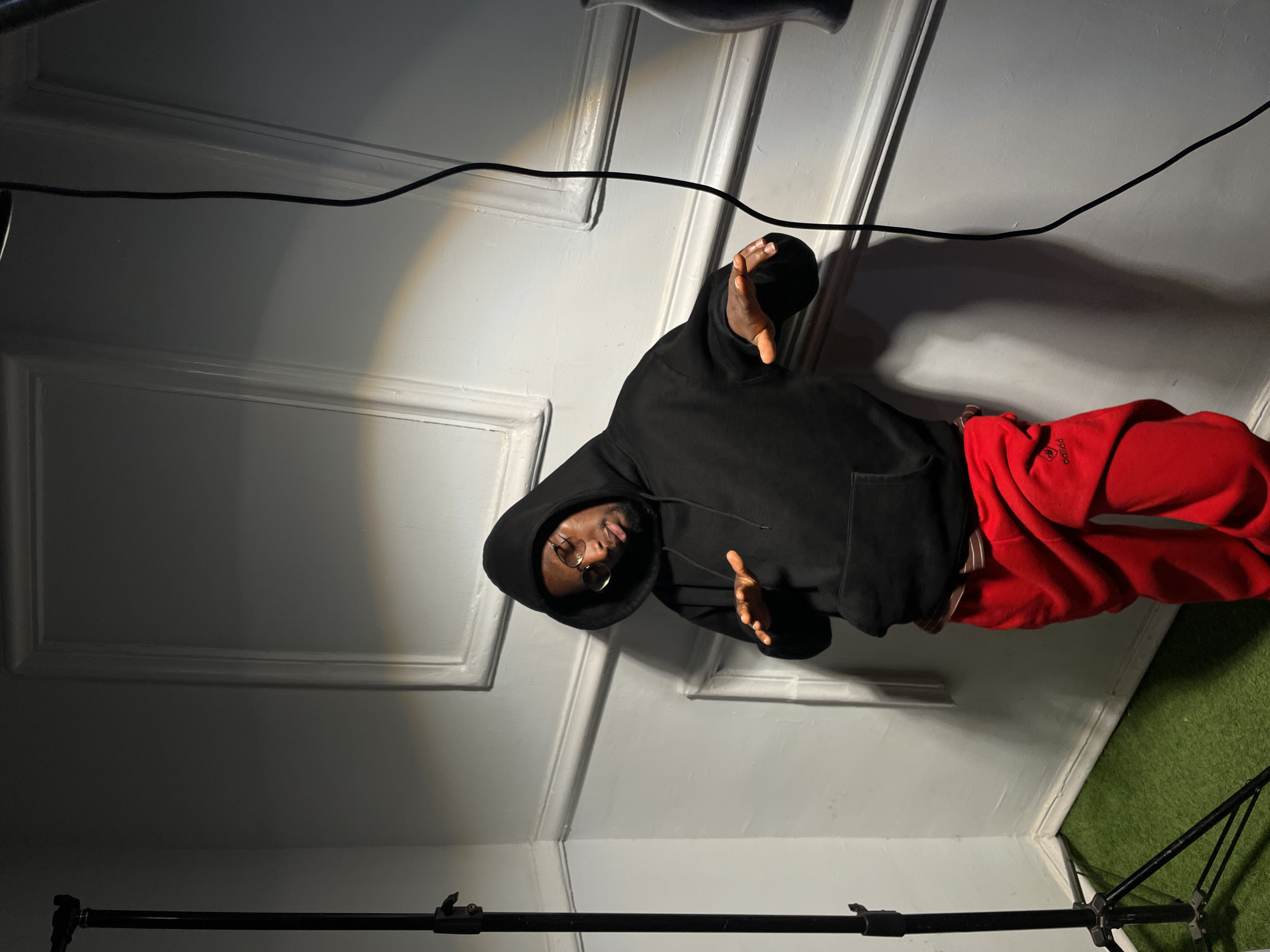 An individual, wearing a black hooded sweatshirt and red track pants, stands in an indoor setting, posing for a photograph. The image is rotated 90 degrees counter-clockwise. The person, likely male, wears round, gold-rimmed glasses and gazes directly at the viewer. Their hands are raised and open, palms facing forward, positioned near their head in a gesturing pose.
The background features light-colored walls with decorative molding. A focused light source illuminates the subject, casting shadows on the wall behind them. A dark cable stretches across the upper background. A black tripod or light stand is visible in the foreground. The floor transitions from a light surface to a section of green artificial turf on the right. Faintly visible on the left leg of the red pants is the word "Adidas". The setting, illuminated by artificial light, appears to be a studio or a prepared room in Barakin Akawo, Nigeria. The time of day is not discernible.