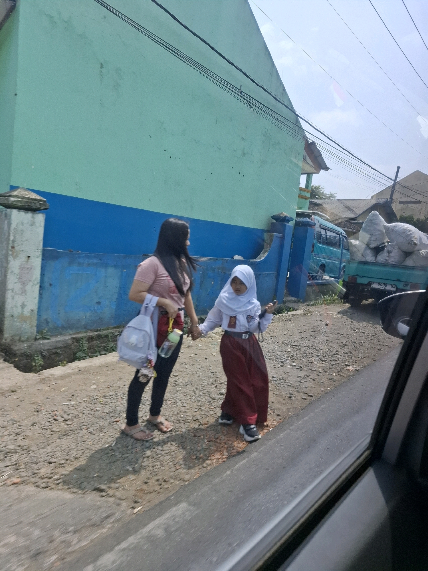 In Lembursawah, Indonesia, a woman in a pink t-shirt and black leggings, carrying a light blue backpack, holds hands with a young girl in a white hijab and red skirt, a school uniform. They walk on a gravelly road beside a turquoise and blue wall. Behind them, a teal pickup truck is loaded with large white sacks. The scene takes place on a sunny day under a hazy sky, with power lines visible overhead.