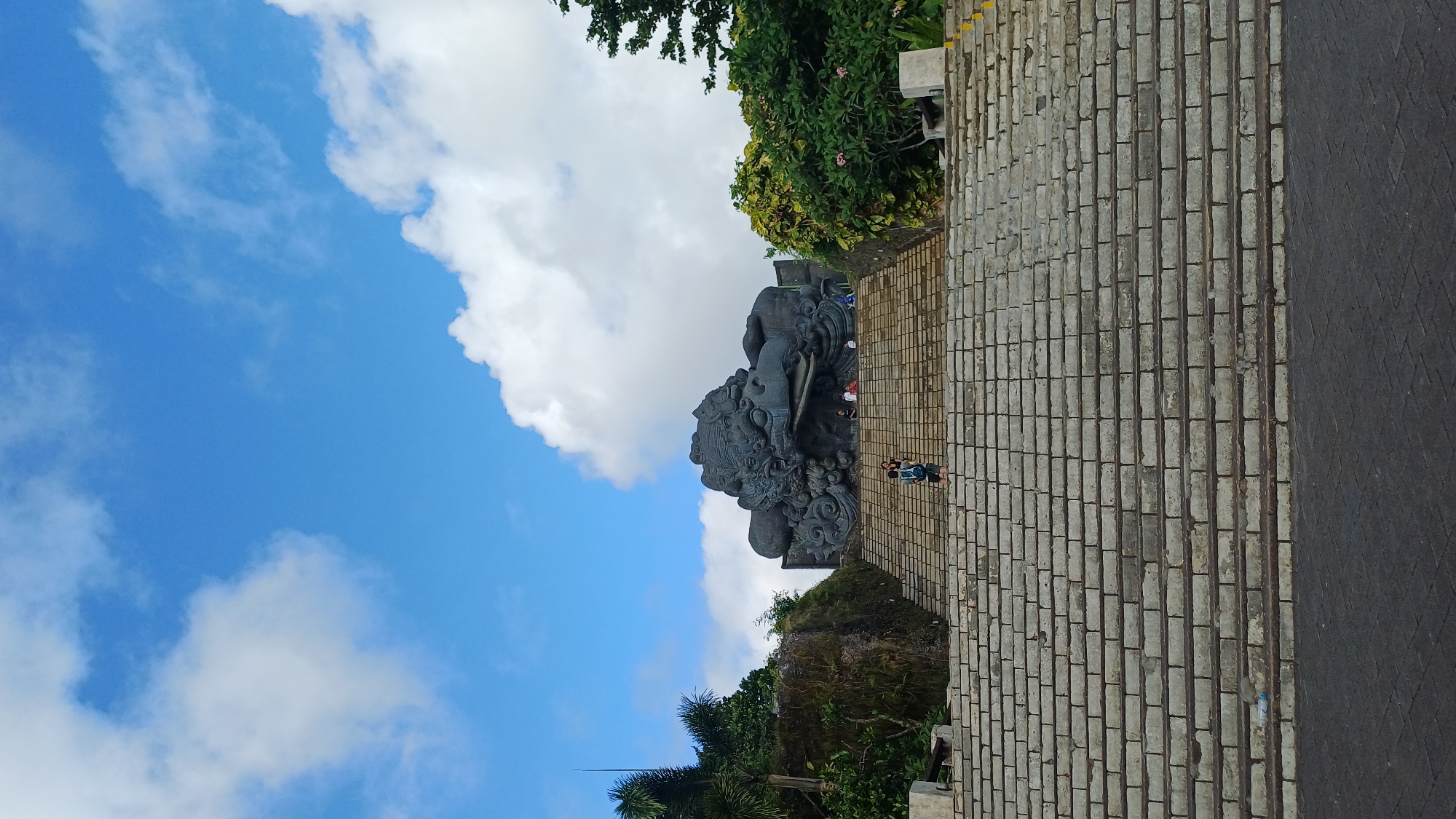 The image shows a large, gray stone sculpture of an elephant-headed figure, likely Ganesha, in Kuta Selatan, Indonesia. The sculpture is placed on a stone platform or ledge, positioned at the edge of a brick-paved walkway or stairway. The sky is visible in the background, with blue sky and white clouds. A few people are seen walking near the sculpture. Vegetation is present on either side of the sculpture and stairs.