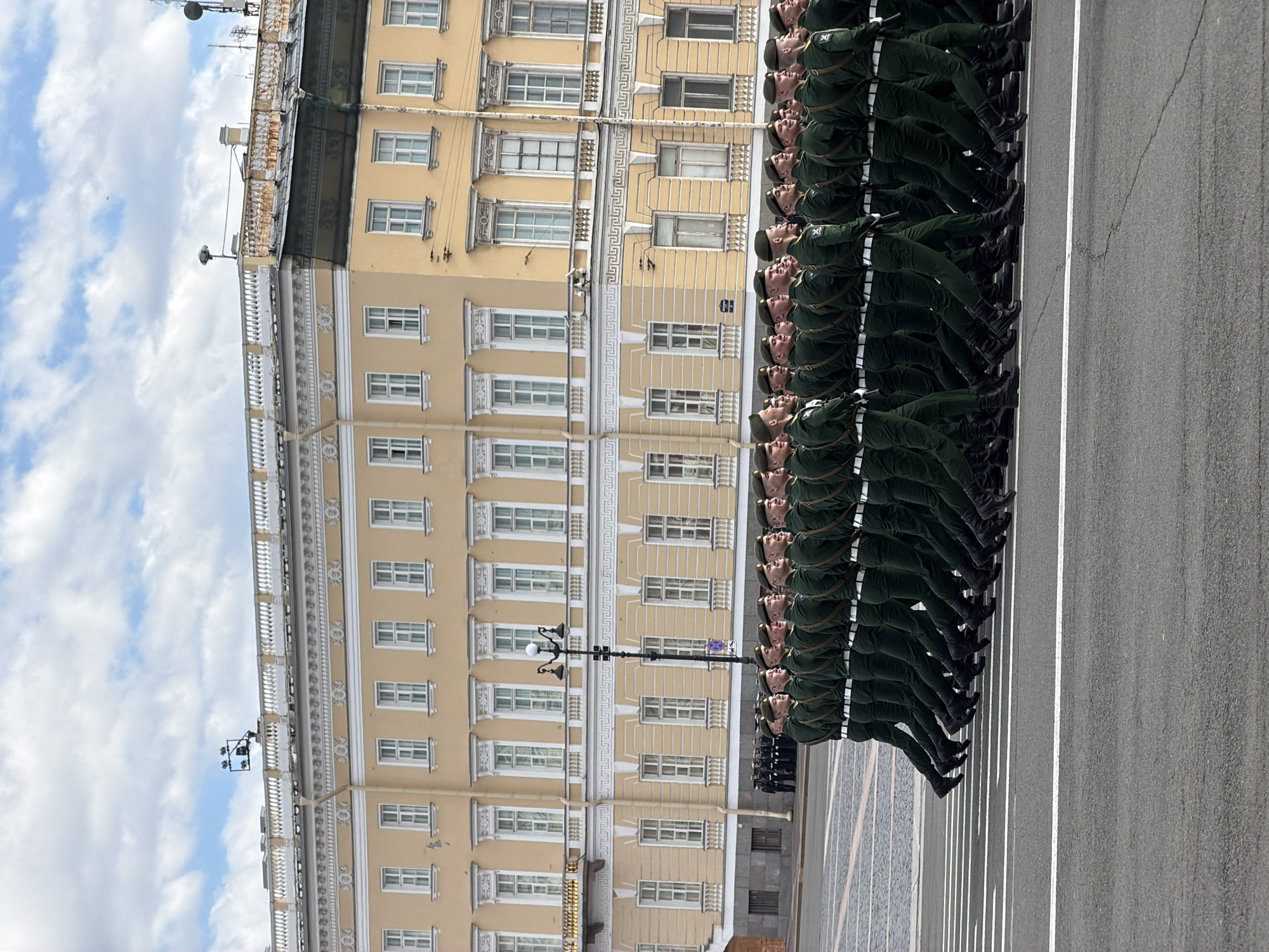 The image shows a military parade in Saint Petersburg, Russia. A large group of soldiers in green uniforms and black boots are marching in formation on a street. The street has white lines painted on it. To the left of the soldiers is a tall, ornate, beige building with multiple rows of windows. The sky is partially cloudy with blue patches. Streetlights are visible along the street.