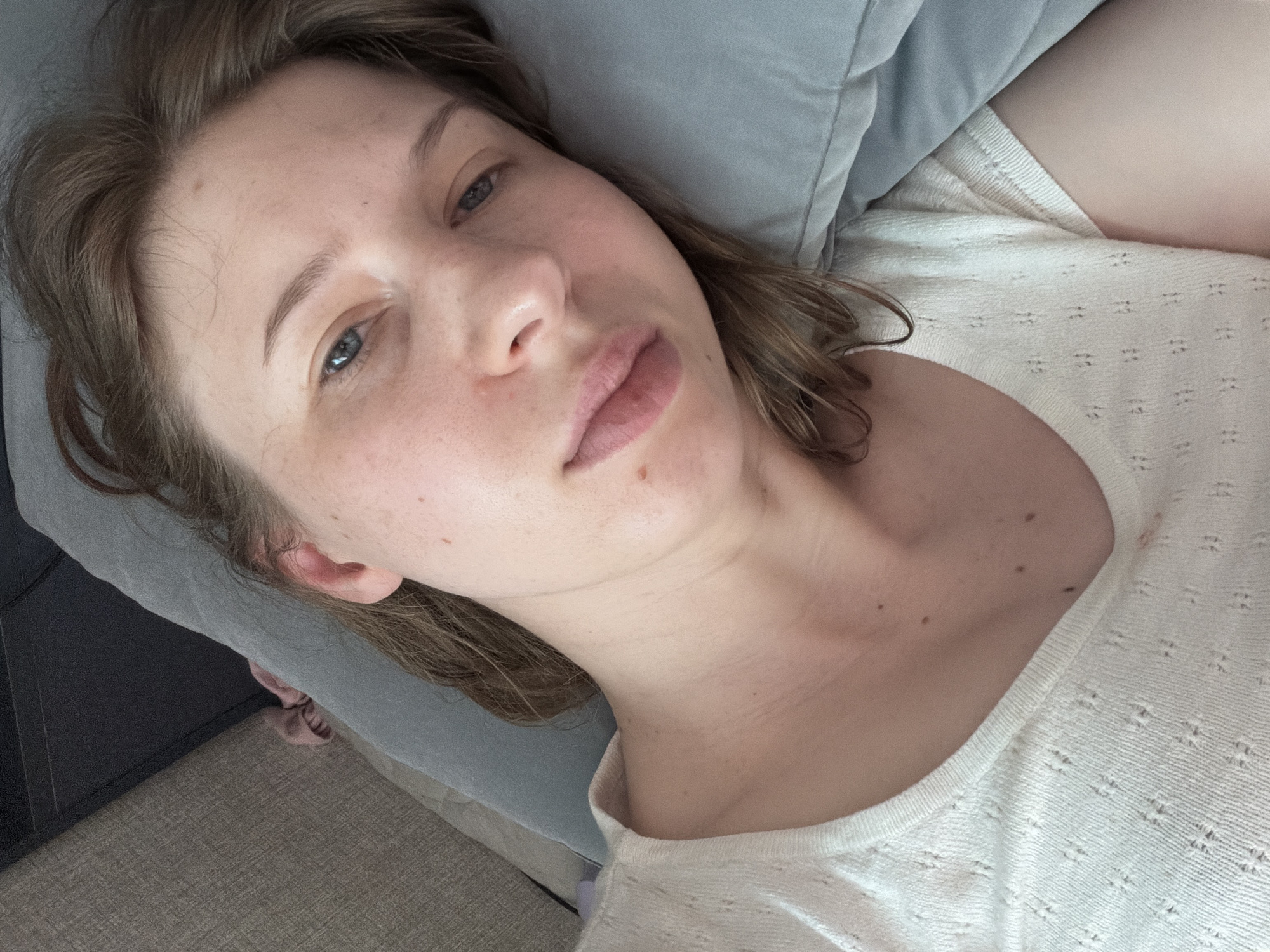 The image shows a close-up, overhead view of a young woman lying in bed, possibly in Dongducheon-si, South Korea. The woman has fair skin with visible freckles and a few moles on her face and neck. Her eyes are open and looking towards the camera, with light blue irises. Her lips are slightly parted. She has medium-length, light brown hair that is loosely styled. She is wearing a light-colored, textured t-shirt with a V-neck. The background consists of a gray pillow and bedding, with a hint of a dark headboard or furniture. The lighting appears to be natural, coming from above and to the side.
