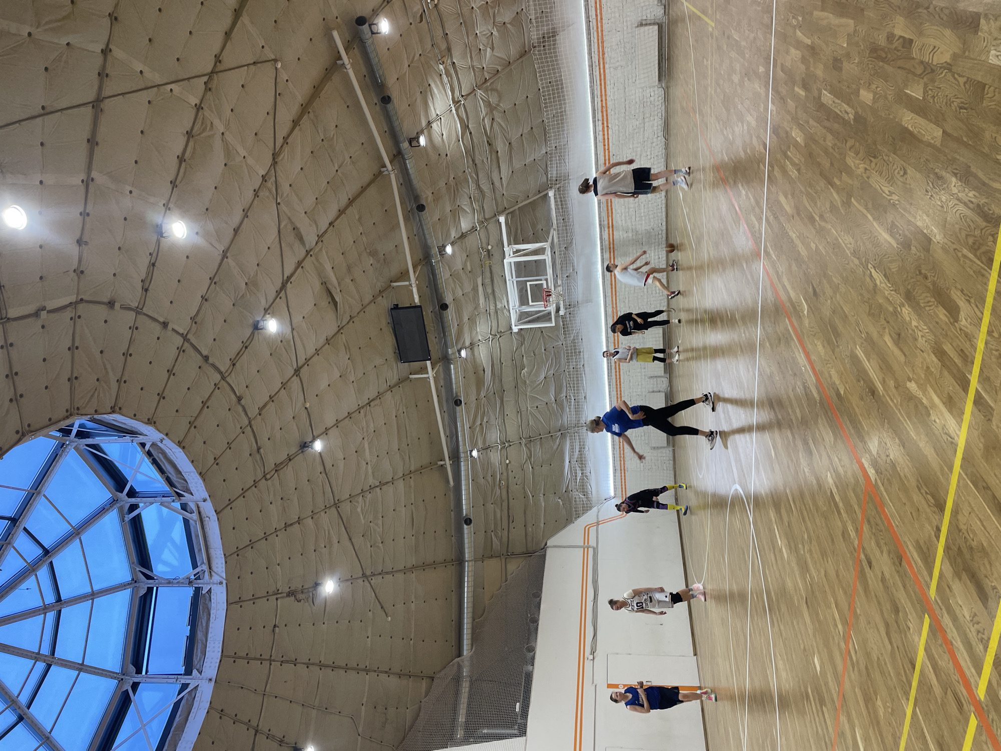 The image shows an indoor basketball court with a high, domed ceiling. The ceiling is covered in a beige, quilted material with regularly spaced lights and a large, circular skylight on the left side. Underneath the ceiling, there is a basketball hoop and a net hanging down. The court itself is made of light-colored wood with yellow and orange lines marking the boundaries and zones. There are several people on the court, appearing to be practicing or playing basketball. They are dressed in athletic wear, including shorts, leggings, and jerseys. They are spaced out along the court, some standing still and others in motion. The image depicts a basketball practice session in what is likely a sports complex or gymnasium in Saint Petersburg, Russia, considering the unique architectural design and typical sports facilities.