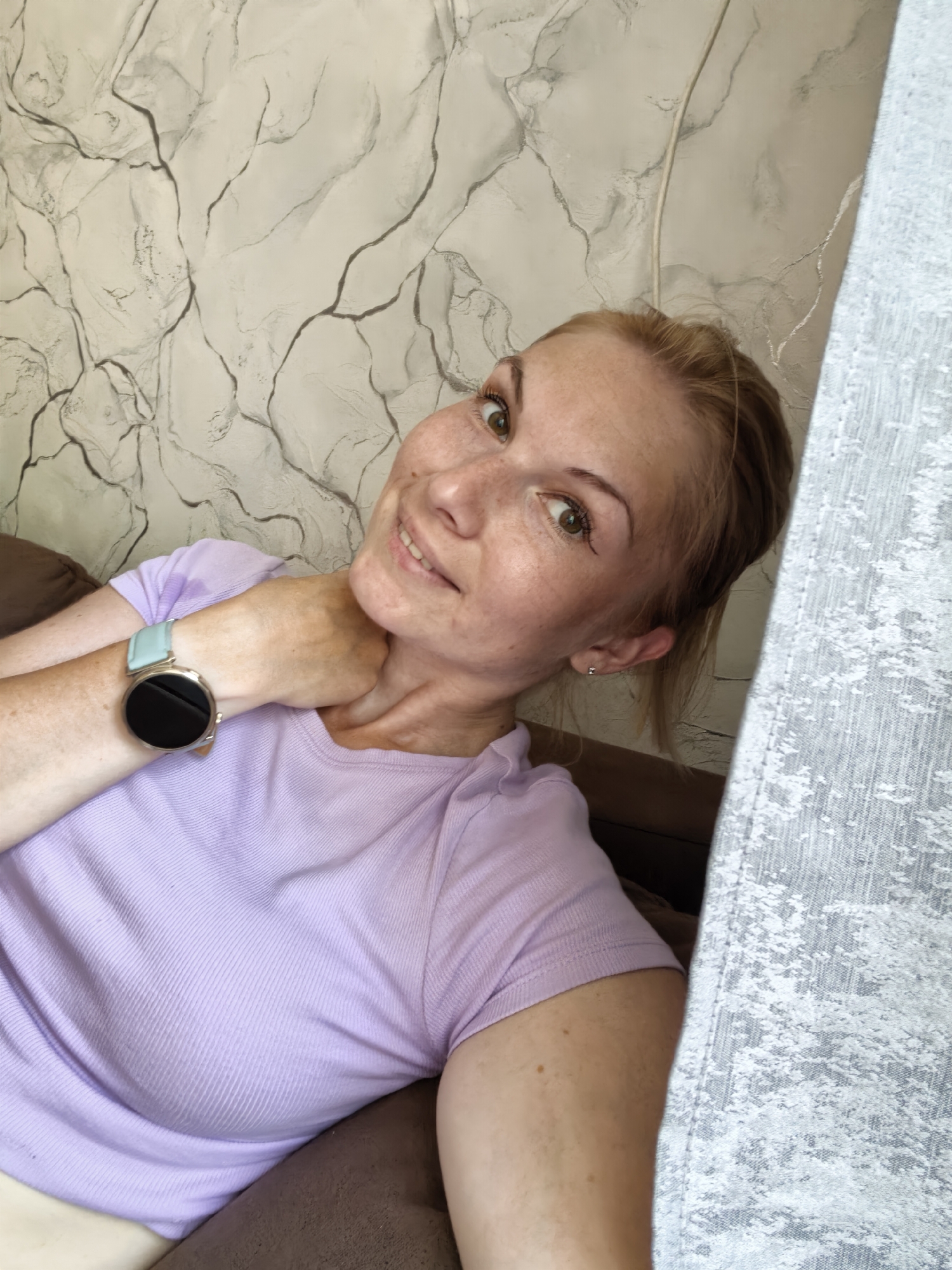 A young Caucasian woman with blonde hair and green eyes is photographed from a high angle, looking up at the camera. She has freckles across her nose and cheeks. She is wearing a light purple t-shirt and a smartwatch with a light blue band on her left wrist. Her right hand is resting on her neck. She is positioned on a brown couch, with a grey and white patterned wallpaper behind her and a grey curtain to her right. The scene appears to be indoors, possibly in a living room. There is no specific indication of the location within the city of Sterlitamak, Russia.
