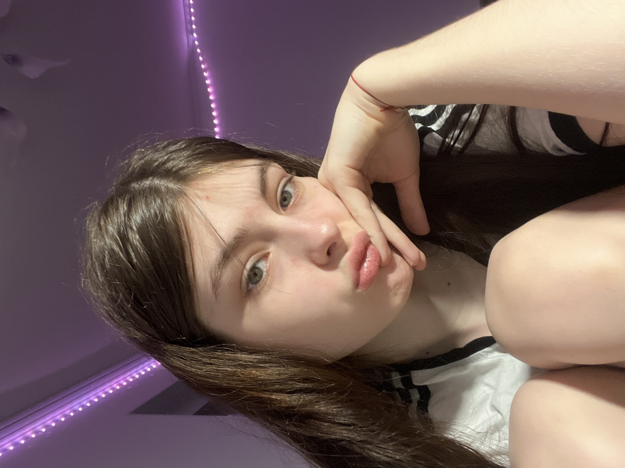 A young woman with long brown hair is positioned in the foreground, her face close to the camera, and with her face tilted and turned slightly left. She is in a pose with pursed lips and her hand resting on her face. She is wearing a white and black striped shirt. The background is dominated by a purple wall illuminated by a string of LED lights. There is no visible evidence in the image indicating the location is the city of Saint Petersburg, Russia.