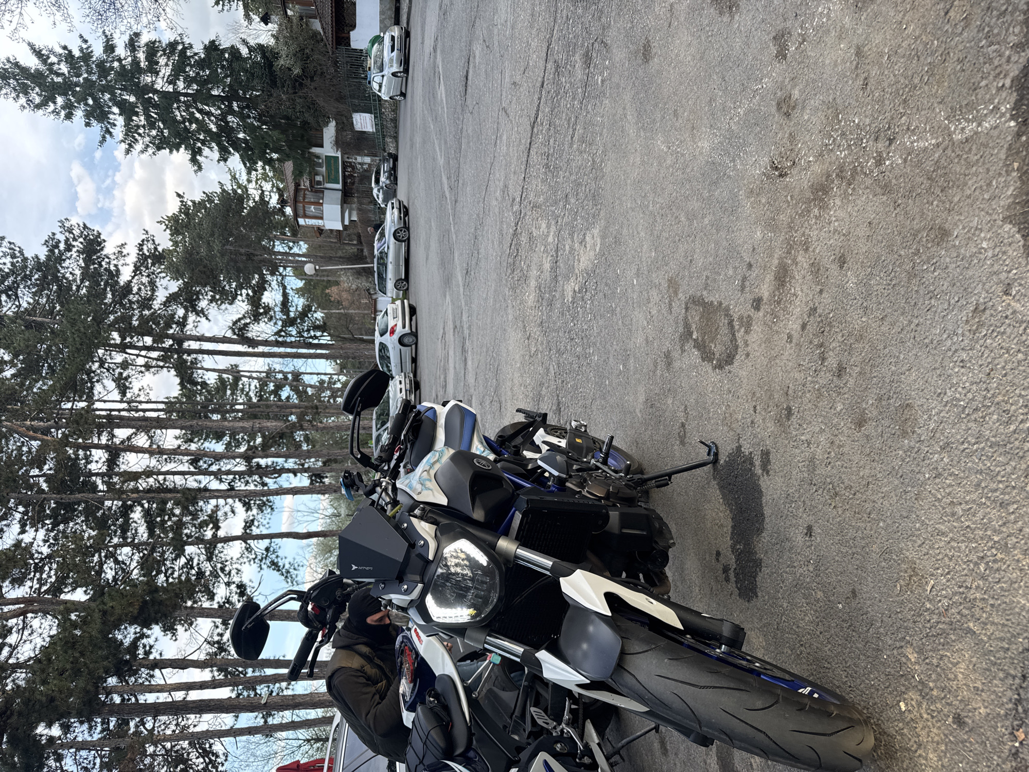 The image shows two motorcycles parked in a paved lot next to a row of trees. The motorcycles are white and blue, with visible front tires, headlights, and handlebars. One motorcycle is closer to the viewer than the other. A person wearing dark clothing, including a jacket and head covering, is near the front motorcycle. Several cars are parked in a row behind the motorcycles. Buildings are visible behind the cars and trees. The sky is visible through the trees.
Based on the available information, I cannot confirm the specific location in the city of Kyustendil, Bulgaria. More evidence would be needed to establish the exact location.