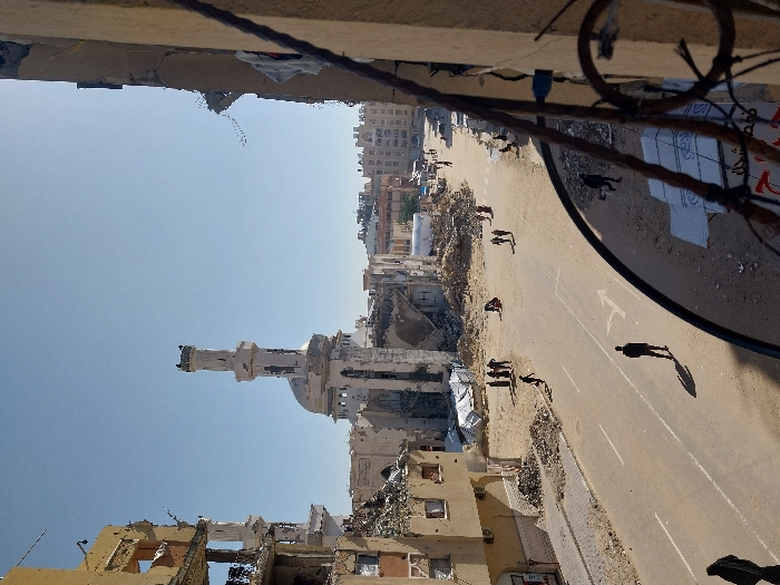 The image depicts a severely damaged urban landscape in Khan Yunis, Palestinian Territory, viewed from an elevated perspective overlooking a street. The scene is dominated by widespread structural destruction under a clear blue sky.
In the mid-ground, the prominent white Al-Farouq Mosque stands with its distinctive minaret and central dome, both showing significant visible damage. To the left, a multi-story building is extensively damaged, with large sections of its facade missing, revealing exposed interiors and rebar. Adjacent to this, and along both sides of the street, are substantial piles of concrete rubble, twisted metal, and other debris, with some white tarpaulins visible amongst the wreckage.
The paved street, running through the center of the image, has faded white lane markings and a white arrow painted on its surface. Approximately 15-20 individuals are present. In the foreground, a lone figure in dark clothing walks on the street, casting a long shadow. Another individual is seen running across the street from left to right in the mid-ground. Several groups of people are scattered across the scene: a group of approximately five stands near the left side of the street amidst the rubble, two individuals walk further down the street on the right, and others are seen walking, standing, or bending over in various areas amidst the debris and on the street. A dark, rusted metal railing or structural element is prominent in the immediate foreground on the right, suggesting the vantage point.