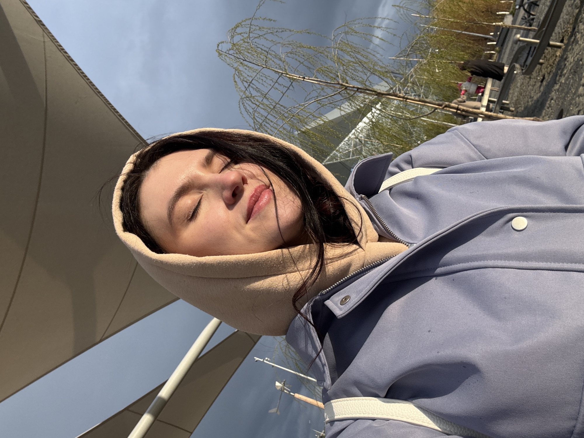 A woman is taking a selfie outdoors. She is wearing a light blue jacket with a beige hoodie underneath. Her eyes are closed, and she has dark hair. The background includes a cloudy sky, a building with a geometric structure, and trees with sparse foliage. Metal structures are visible, along with some indistinct figures in the distance. The location is Kazan, Russia.