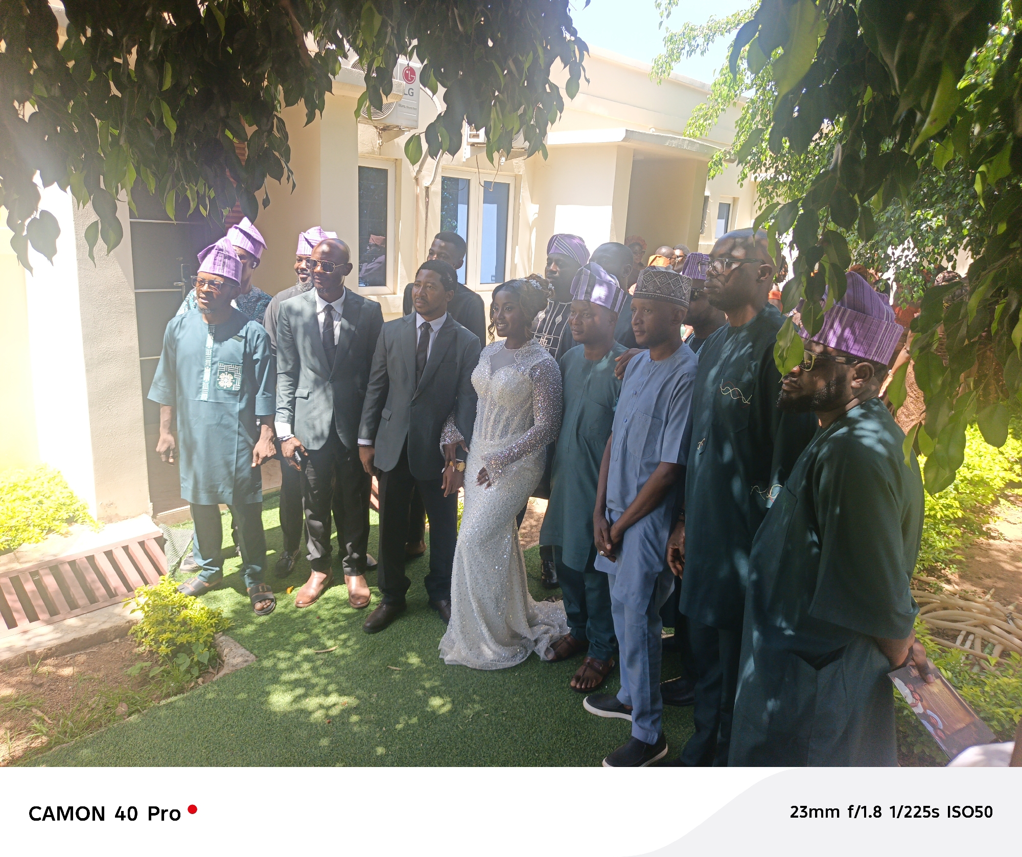 A daytime outdoor photograph shows a group of people posing for a photo. The group includes men in suits, traditional African clothing and headwear, and a woman in a sparkling white dress. They are positioned in front of a light beige or cream-colored building, which appears to be a residence, with an air conditioning unit mounted on the exterior wall. The location may be in or near Barakin Lerawi, Nigeria.
The ground is covered with artificial turf, and there is vegetation around the group. Some of the men in the group are wearing glasses, and others are clean-shaven. The lighting is bright and sunny, with some glare affecting the image.