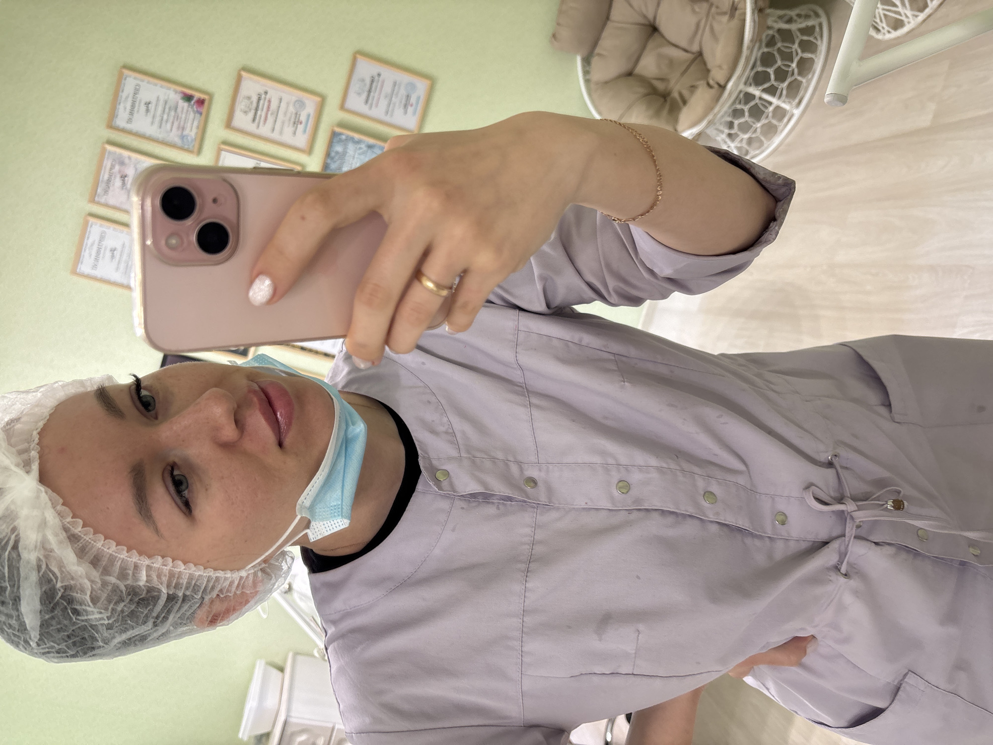 A woman in a light purple medical coat, hairnet, and blue surgical mask is taking a selfie with a pink smartphone. The setting appears to be a doctor's office. The woman is wearing a gold bracelet and a gold ring. In the background, there are framed certificates on the wall and a white chair with a beige cushion. The location is Kostoryata, Russia.