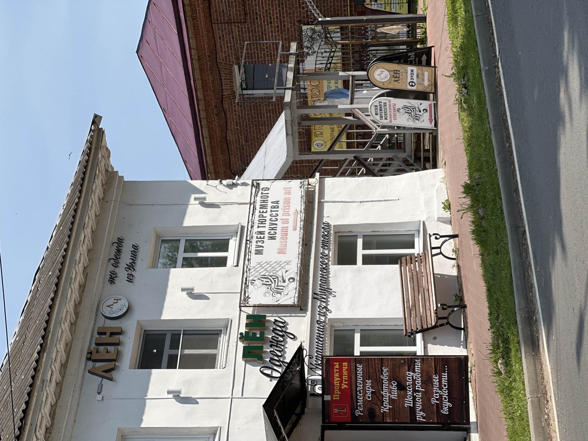This is a street-level, eye-level, medium shot of a white, two-story building with a dark grey, corrugated metal roof in Selivanovo, Russia. The building has several windows, and signage is displayed prominently on its facade.
To the left, on the upper part of the building, is a sign that reads "ЛЁН" in large, three-dimensional gold letters, followed by a circular logo with stylized plants and the letters "ЛЁ". To the right of this logo, in a script font, is the text "эко одежда из Углича". Below this, a white sign with black text reads "МУЗЕЙ ТЮРЕМНОГО ИСКУССТВА Museum of prison art".
A dark awning extends from the building to the left, and below it is a vertical sign with a red anchor symbol and the text "Продукты Углича". Below this, on a dark wooden plank sign, are listed "Ремесленные сыры", "Крафтовое пиво", "Шоколад ручной работы", and "Разные вкусности...".
To the right of the white building, a brick building with a dark red, sloped roof is visible. Adjacent to the white building's facade, there is an outdoor staircase with metal railings leading up to the second floor. Several A-frame signs are placed on the sidewalk in front of this staircase. One sign features a drawing of a person in striped clothing and text that includes "МУЗЕЙ ТЮРЕМНОГО ИСКУССТВА" and "СУВЕНИРЫ". Another sign, with a stylized "ЛЁ" logo, has text that includes "ЛЁН эко одежда" and "2 ЭТАЖ".
A wooden bench with black metal legs sits on the red brick sidewalk in front of the white building. A grey concrete curb borders the sidewalk, and a paved road is visible in the foreground. The sky is clear and blue. There are no people clearly visible in the frame.