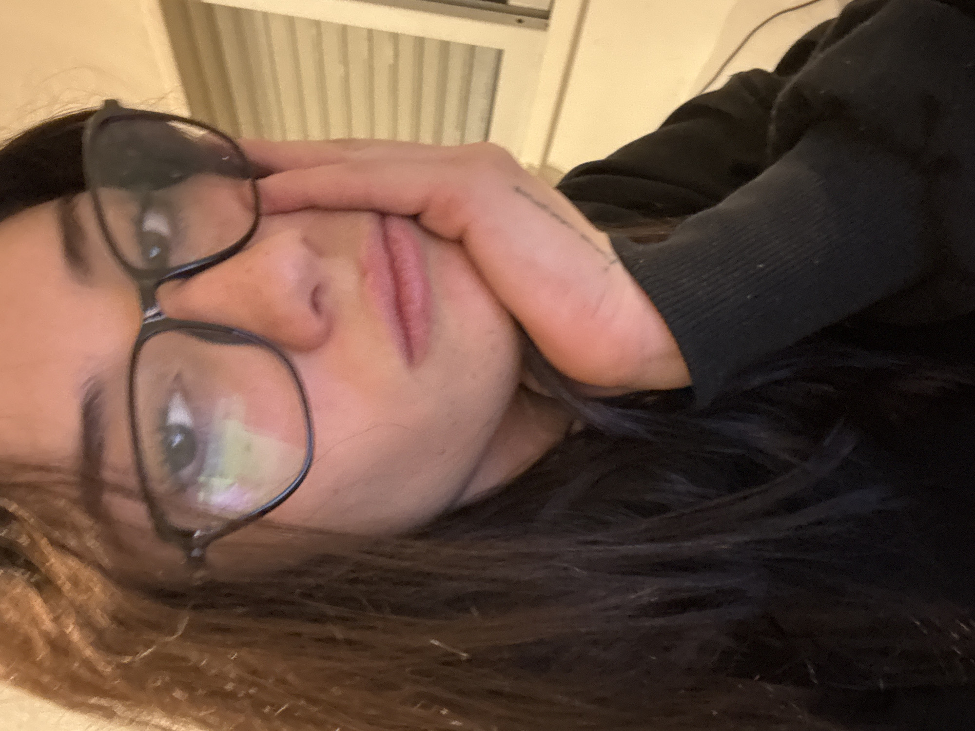 A close-up, angled selfie captures a person with light skin, dark hair, and dark-framed glasses. Their hand partially covers their face near the mouth and nose. They wear a dark-colored sweater or sweatshirt. The background features a blurry, light-colored surface with vertical lines. The location is not discernible from the image.
