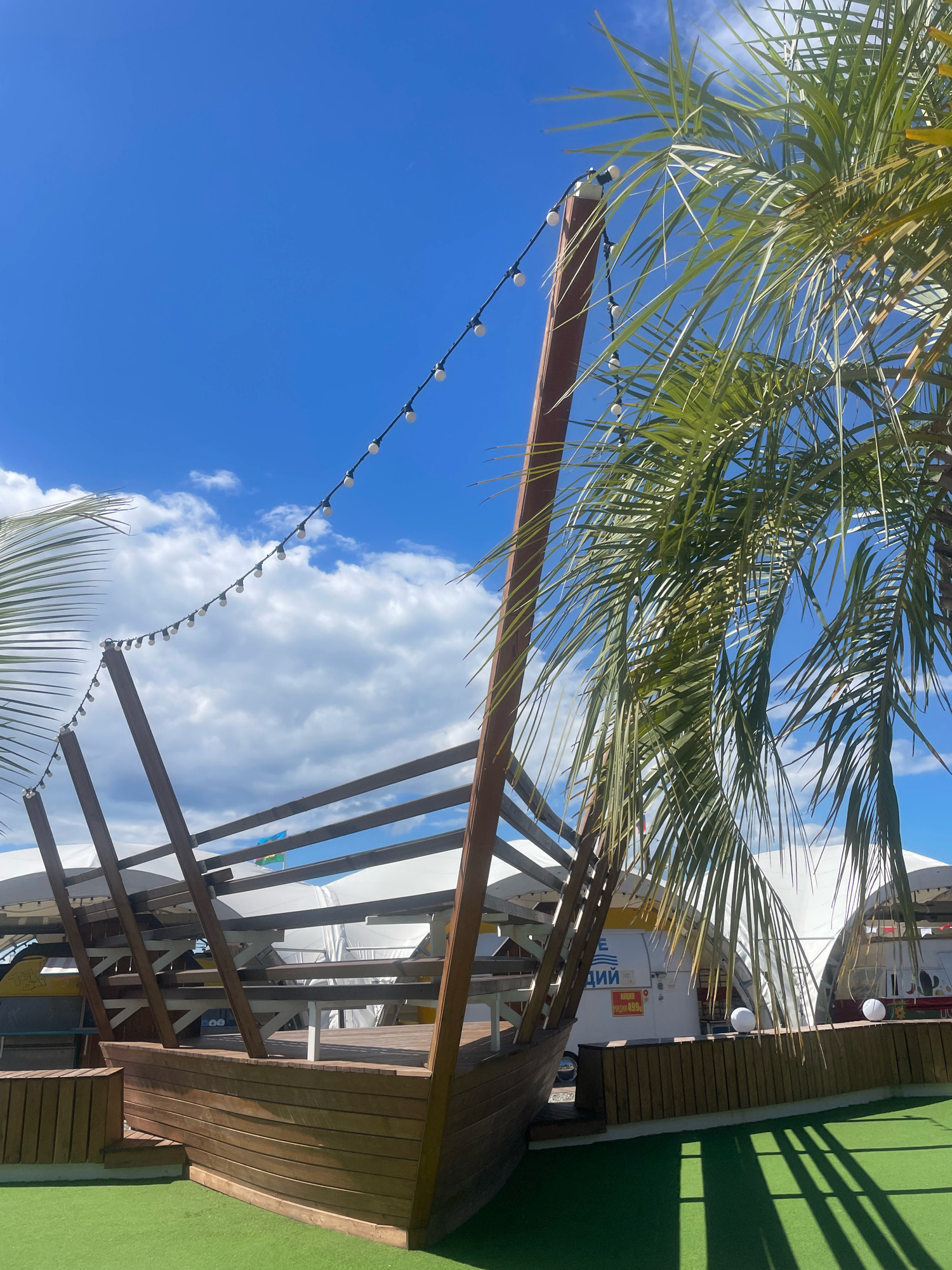The image depicts an outdoor seating area with a wooden structure shaped like the front of a ship. The structure is situated on a bright green artificial turf surface. In the background, there are white tent-like structures and palm trees, suggesting a warm, possibly tropical or coastal environment. A string of white lights hangs above the wooden ship structure, and a clear blue sky with scattered white clouds is visible. A flag is also seen flying in the distance. The overall impression is that of a themed outdoor dining or recreational space. The location in Sochi, Russia is suggested by the context of the request.