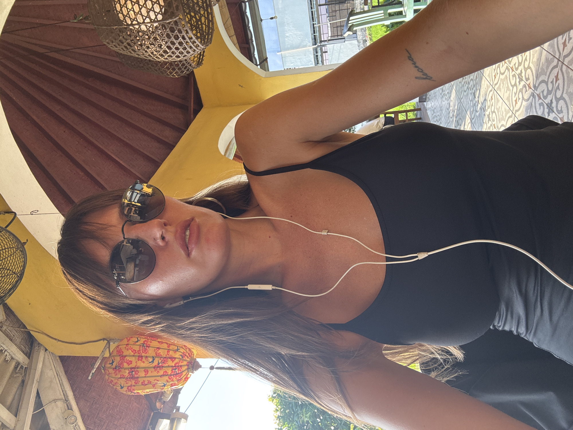 A medium shot captures a woman with long brown hair and bangs, wearing round sunglasses and a black tank top. She has white earbuds in her ears, with the cord running down her chest. A tattoo is visible on her right forearm. She is seated outdoors, with a yellow structure visible behind her. Hanging from the structure are two decorative lanterns, one red and yellow with floral patterns, and another woven. The background also shows glimpses of greenery and outdoor seating. The scene appears to be set in a cafe or restaurant with an open-air setting, potentially in a tropical location like North Kuta, Indonesia, given the style of the lanterns and foliage.