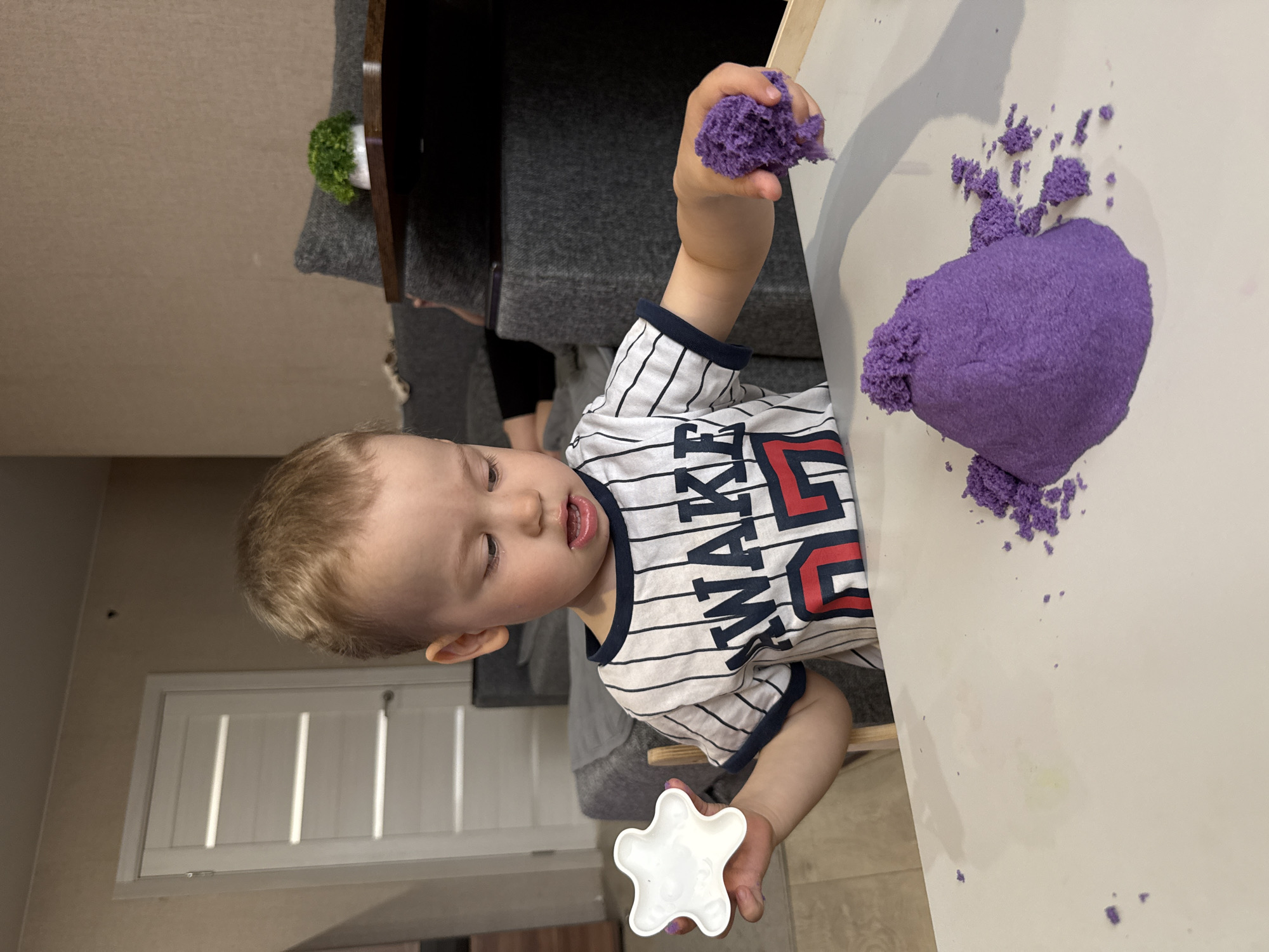 A young child with fair hair is playing with purple kinetic sand on a white table. The child is wearing a white T-shirt with black stripes and a red and blue graphic. The child holds a clump of sand in their right hand and a white star-shaped mold in their left. Additional clumps of the purple sand are on the table around the child. The setting appears to be indoors, with a gray couch and a small potted plant visible in the background. The location is Rusanovo, Russia.