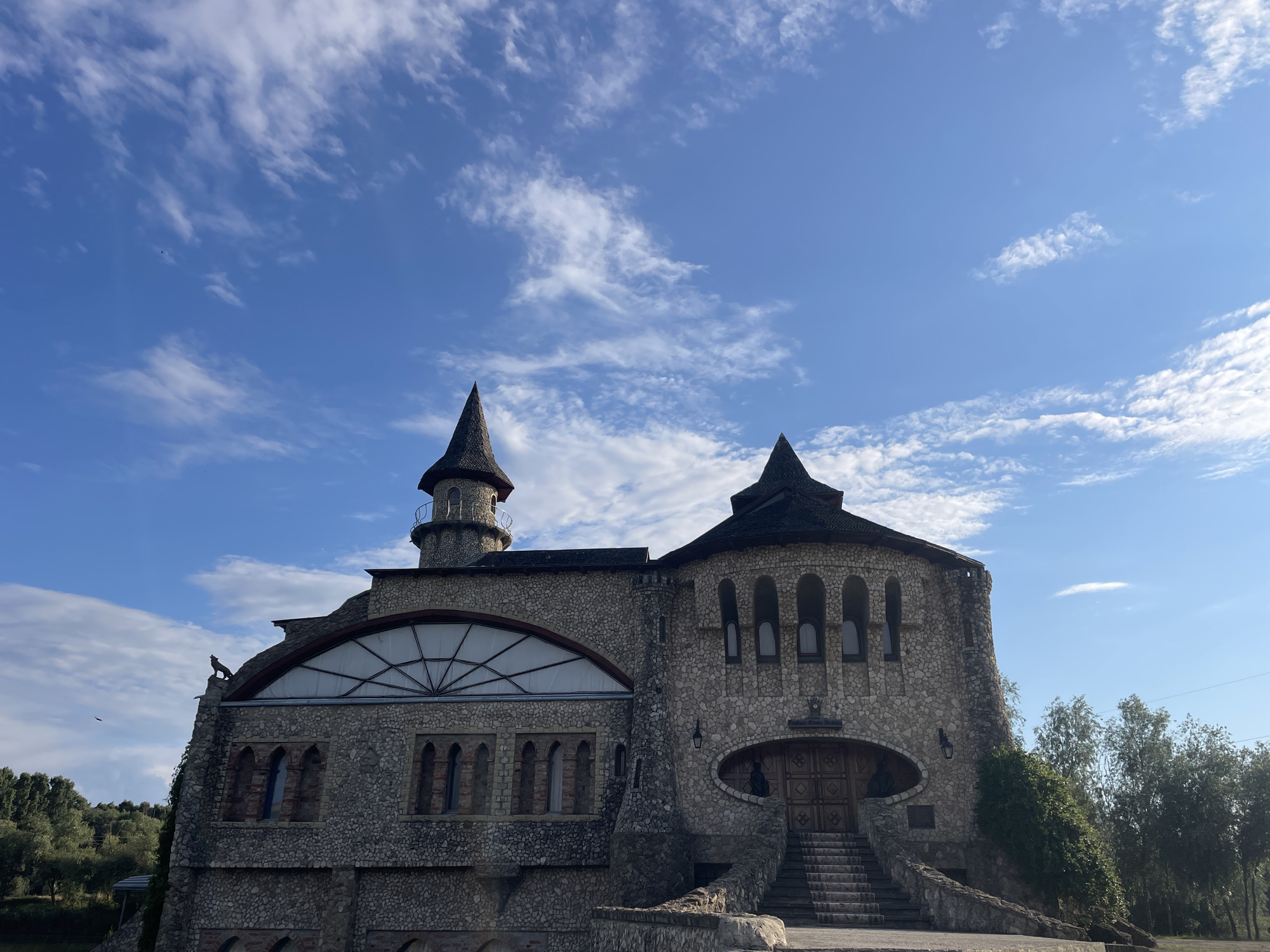 The image shows a unique, castle-like building constructed primarily of stone with conical roofs and turrets. The building has several arched windows and a large arched window with a geometric design on the left side. A statue of a bird of prey is perched on the edge of the roof on the far left. A set of stone stairs leads up to a large wooden door. The sky is blue with scattered white clouds. Trees and foliage are visible to the right and in the background. This building is identified as being located in Colibași, Moldova. No people are visible in the image.