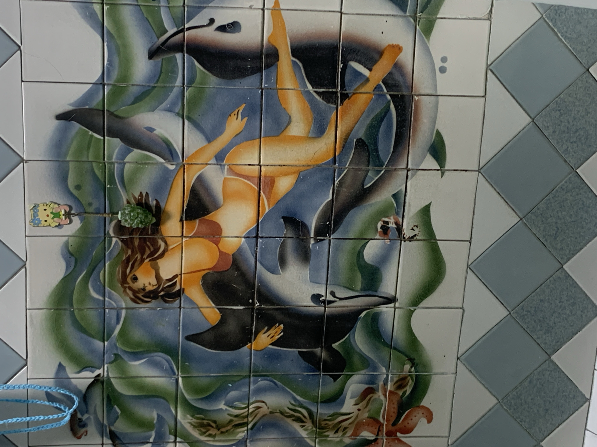 The image depicts a tiled mural featuring a woman in a red bikini swimming with dolphins amidst stylized blue and green waves. The woman has long brown hair and is positioned horizontally, with one arm extended and the other around a dolphin. Two dolphins are visible, one in the foreground being embraced by the woman, and another partially visible in the background. The mural is rendered in a vibrant, illustrative style. Above the mural, a blue beaded cord is partially visible, and to the right, a small decorative item resembling a cartoon character is attached. The mural is bordered at the bottom by a pattern of light blue and white diamond-shaped tiles. There is no visible context that would suggest the mural's location within the city of Tashkent, Uzbekistan.