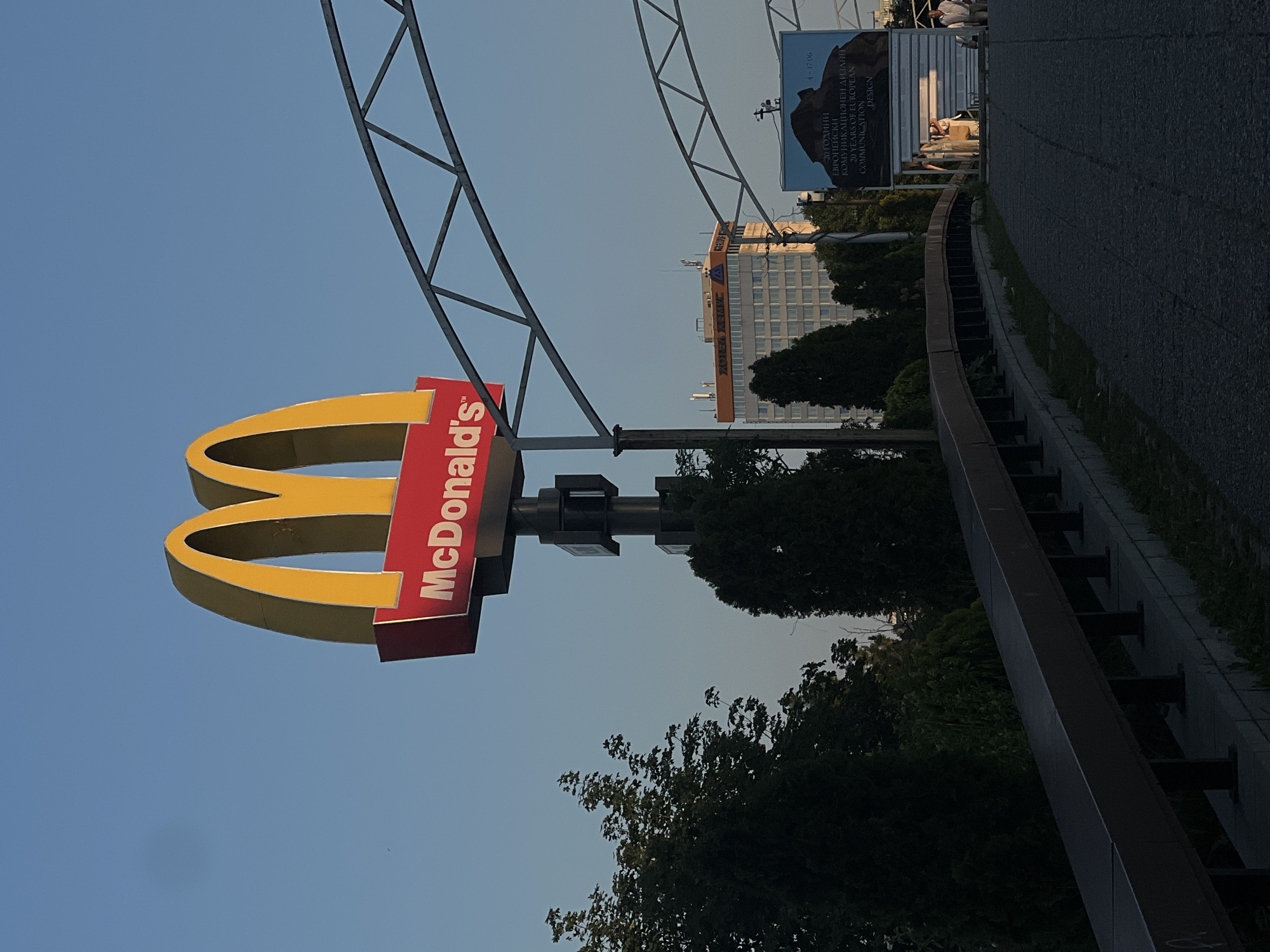 A McDonald's sign with golden arches and a red "McDonald's" logo is prominently displayed against a clear blue sky. The sign is mounted on a tall black pole. In the background, a modern office building with many windows is visible, along with trees and a billboard. A road or pathway is visible in the foreground, leading towards the billboard where a few people are standing. The scene appears to be in an urban setting during the daytime, possibly late afternoon given the lighting.