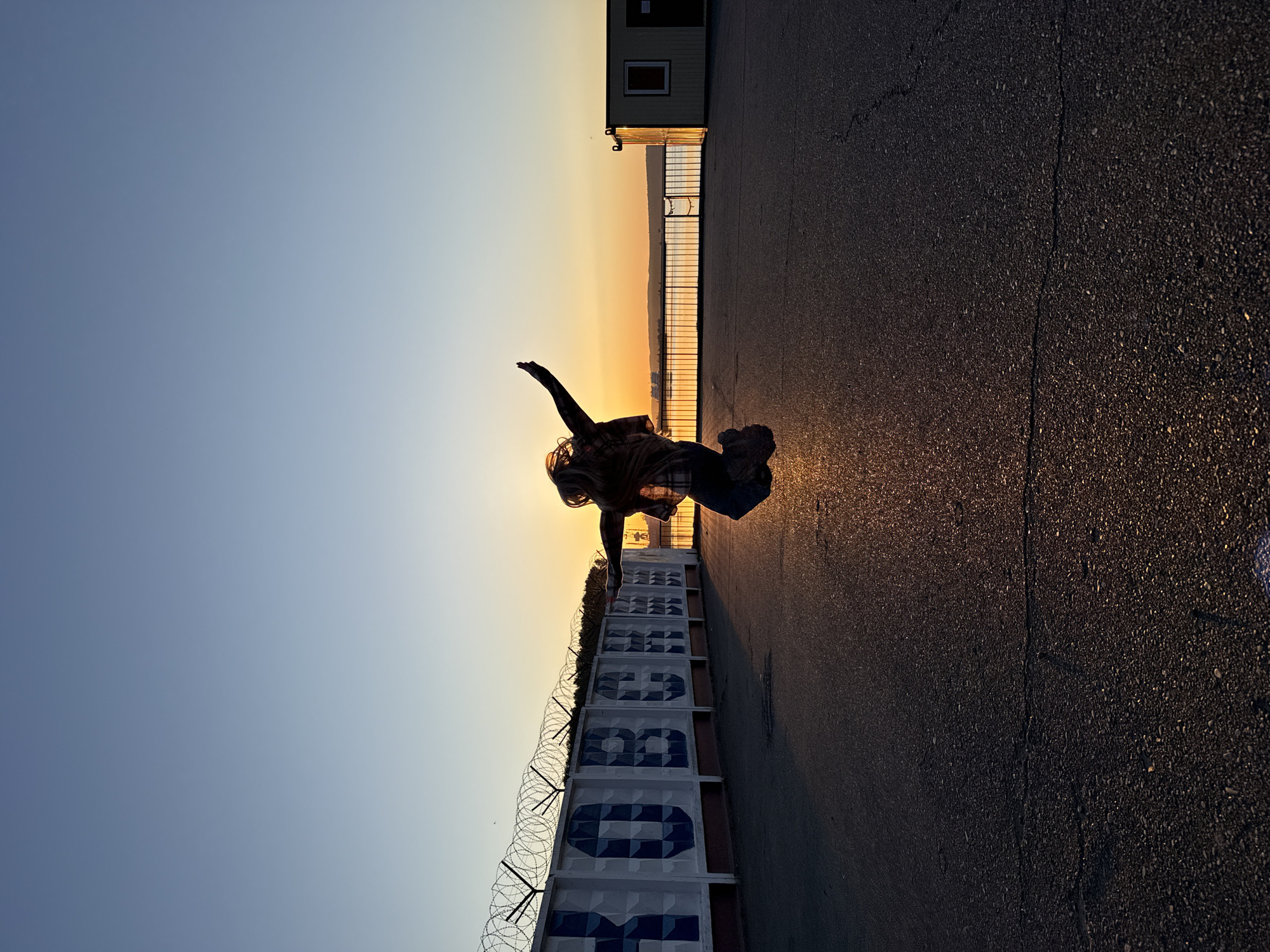The image depicts a person in silhouette against a sunset in Chaikovsky, Russia. The person is positioned between a concrete wall with numbers and razor wire and a paved surface that appears to be a road or walkway. A building with a window is visible in the background on the right. The sky transitions from blue to orange and yellow as the sun sets. The number wall may represent a former factory or other industrial complex.