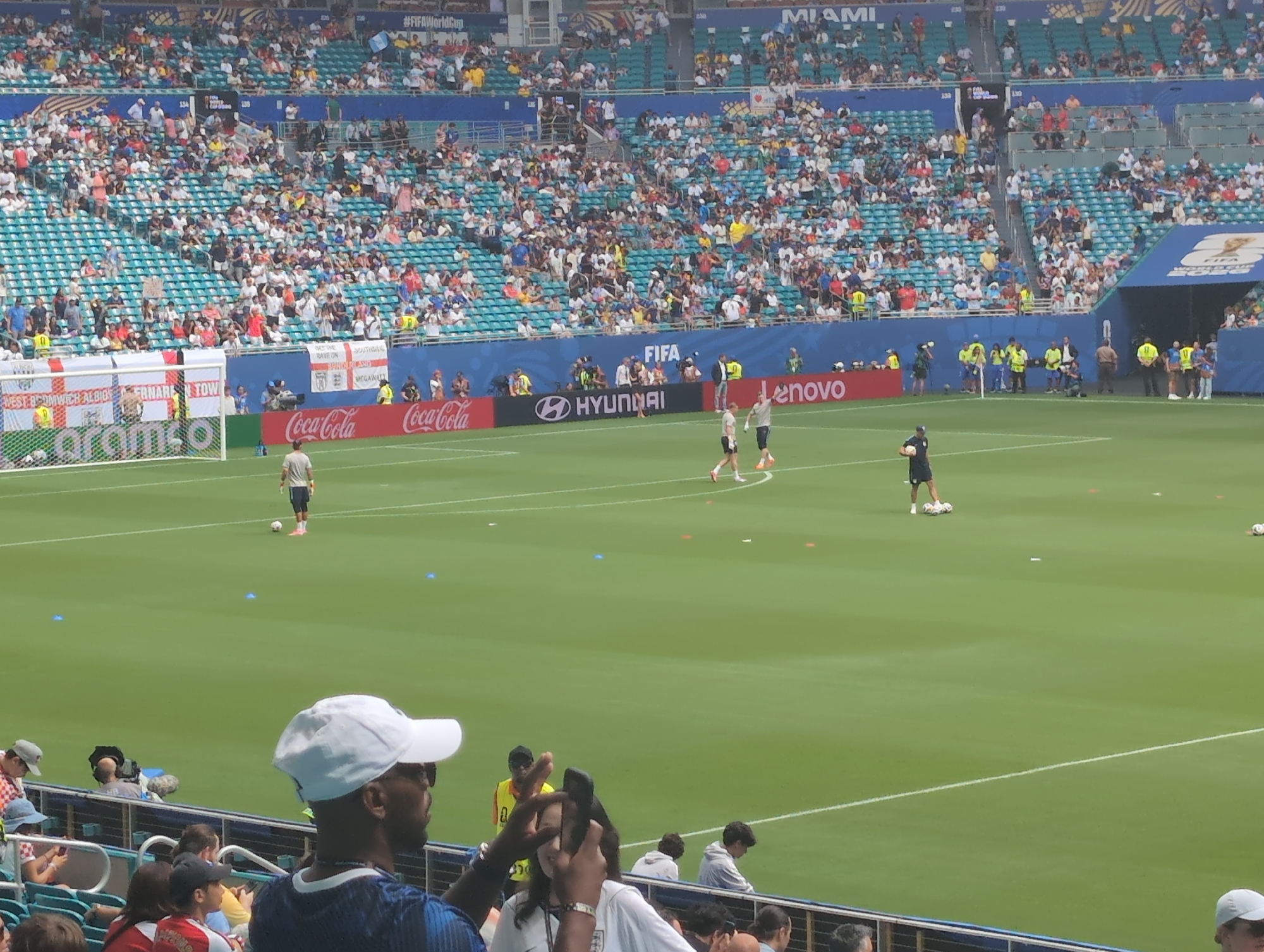 This image captures a wide-angle view of a soccer stadium, likely Hard Rock Stadium in Miami Gardens, Florida, during a match or practice. The stadium is filled with spectators seated in tiered bleachers. The green soccer field is visible, with white lines marking boundaries and a soccer goal on the left. Several people are on the field, including players in soccer attire and individuals in vests that might indicate staff or security. There are also various advertising boards and banners around the stadium, displaying logos from brands like Coca-Cola, Hyundai, and Lenovo, along with FIFA branding, suggesting an official event, possibly related to the FIFA World Cup. In the foreground, several spectators are visible, some holding up phones, indicating they are taking pictures or videos of the event. The overall atmosphere appears to be that of a live sporting event with a significant crowd.